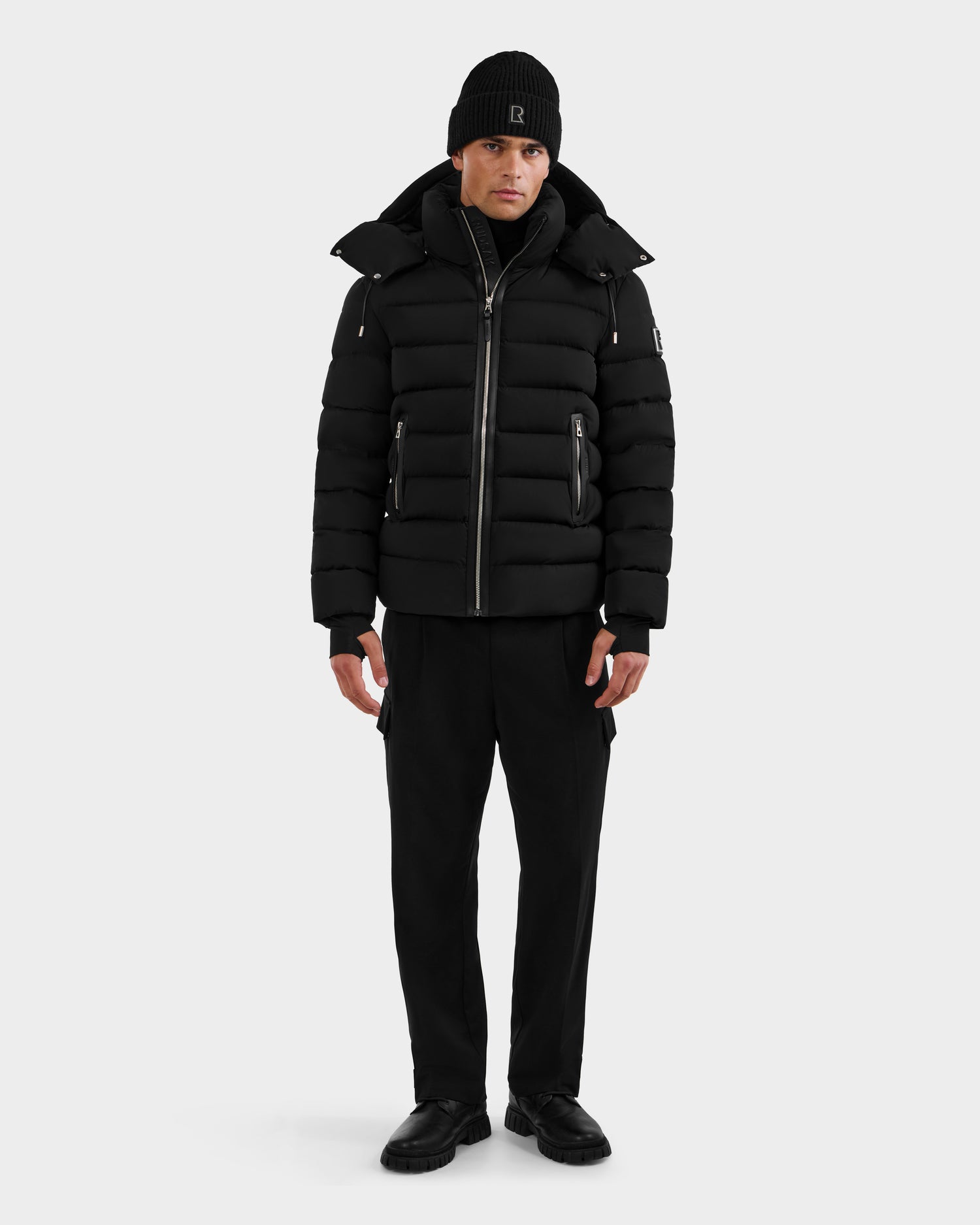 A person faces forward, wearing the Rudsak NAVAN - 6125598 Black men’s quilted down puffer jacket with zipper and pocket details, along with a black beanie, pants, and shoes, against a plain light background.