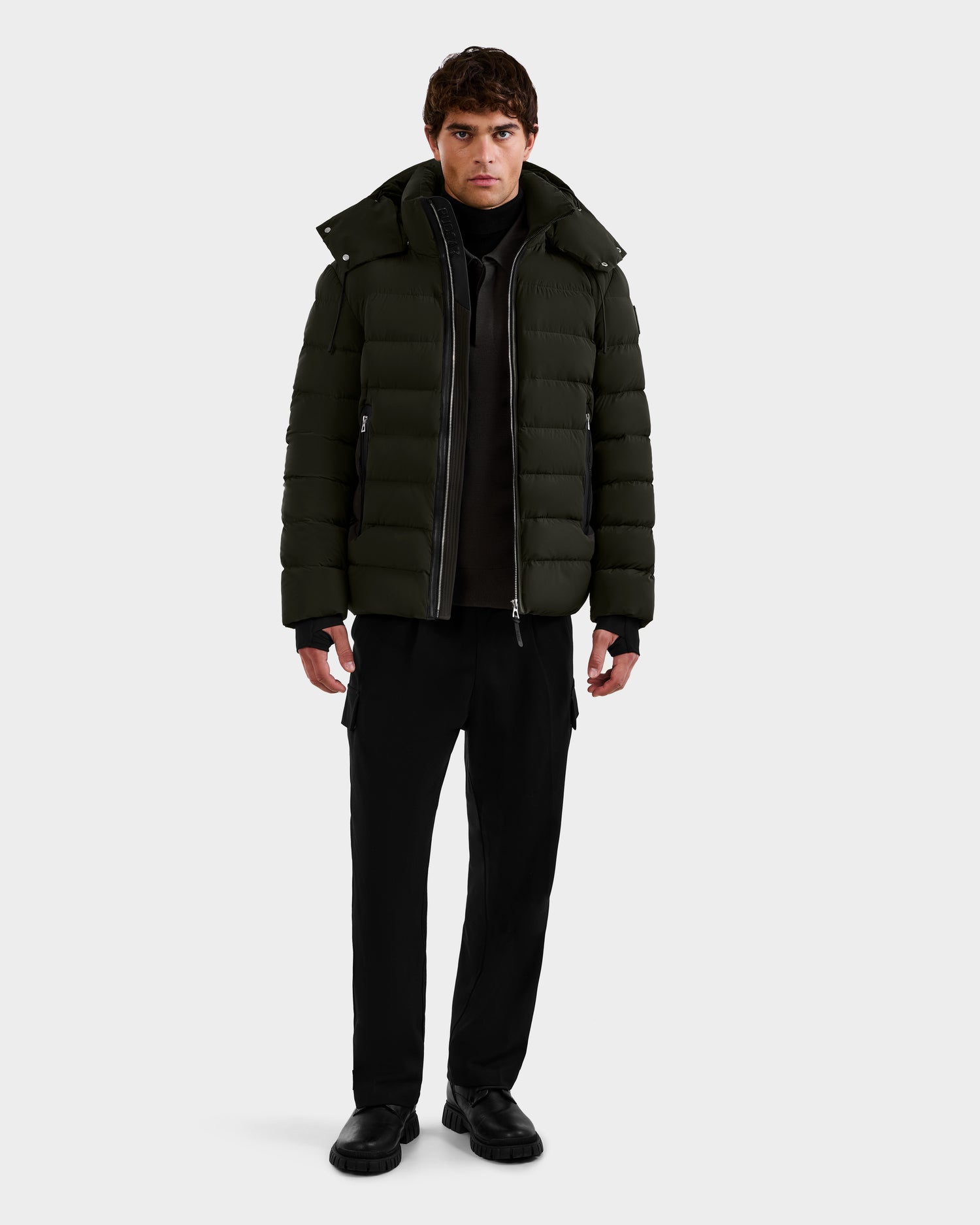 A man faces forward, wearing the Rudsak NAVAN - 6125598 Dark Olive men's down puffer jacket over a black turtleneck with black pants and shoes, posed against a plain white background.