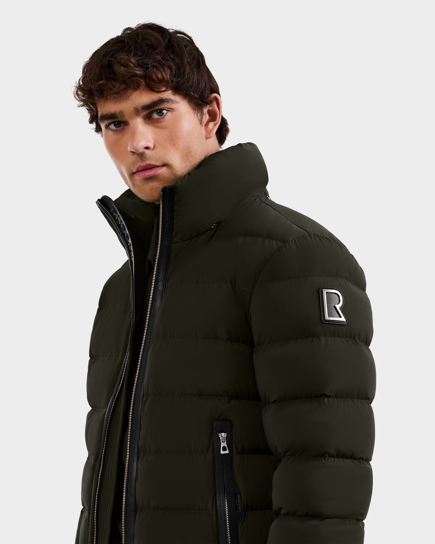 A young man with curly brown hair wears the Rudsak NAVAN - 6125598 down puffer in dark olive, featuring a high collar, visible zippers, and an R patch on the left sleeve. He faces the camera with a serious expression.