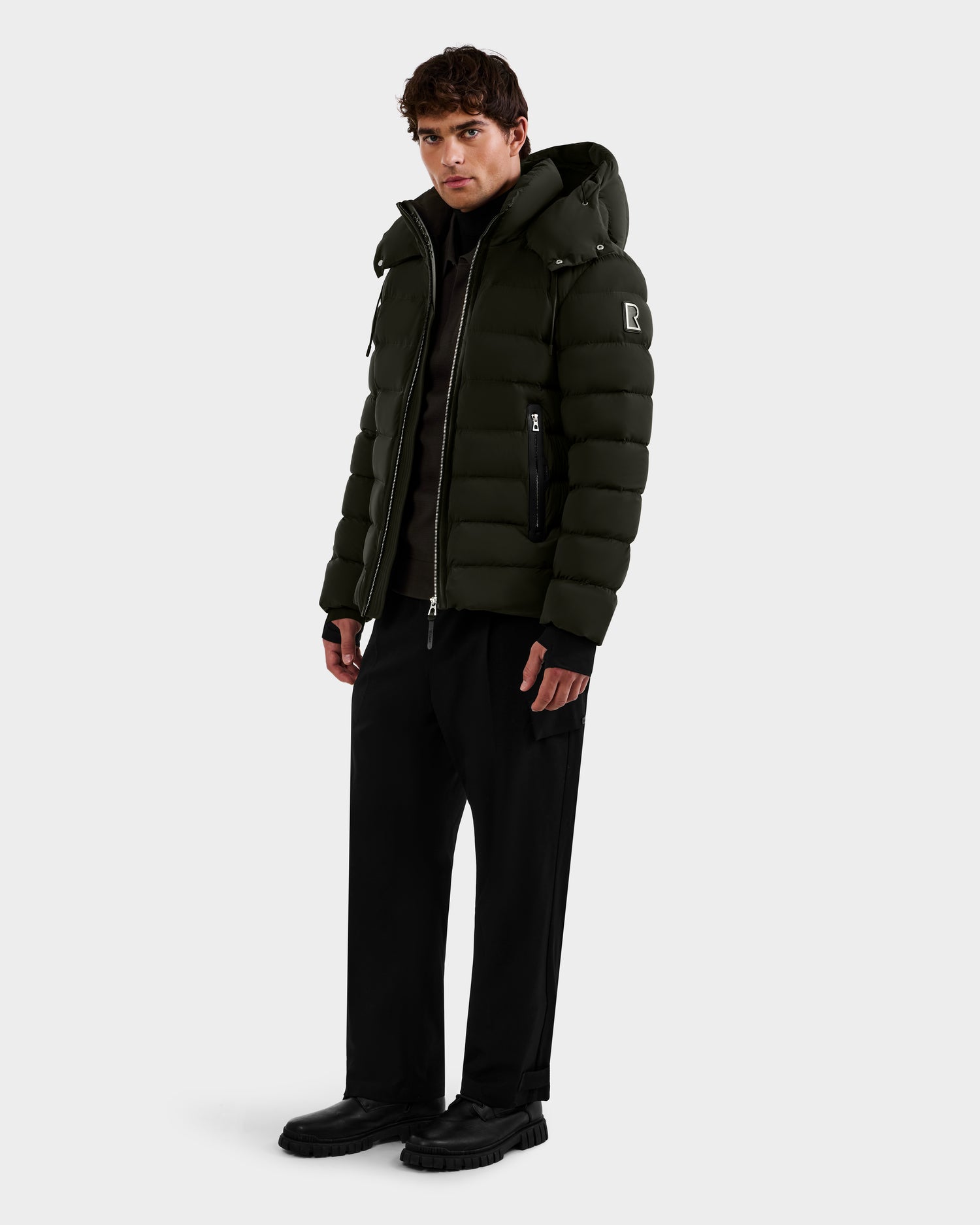 A man faces forward wearing the Rudsak NAVAN - 6125598 Dark Olive quilted puffer jacket with a hood, paired with black pants and shoes, set against a plain light background.