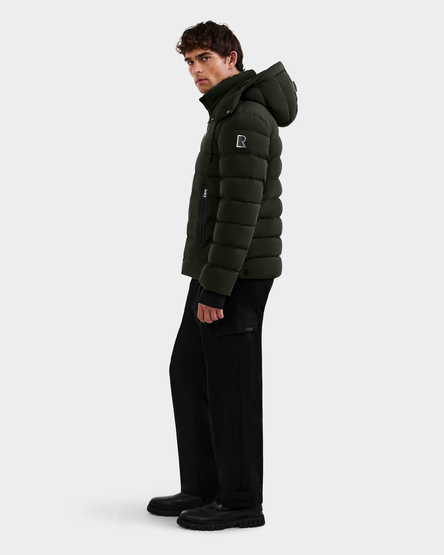 A man stands sideways against a plain light background, wearing the Rudsak NAVAN - 6125598 dark olive quilted puffer jacket with a hood, black pants, and black boots. He looks ahead neutrally, highlighting this stylish men's outerwear.