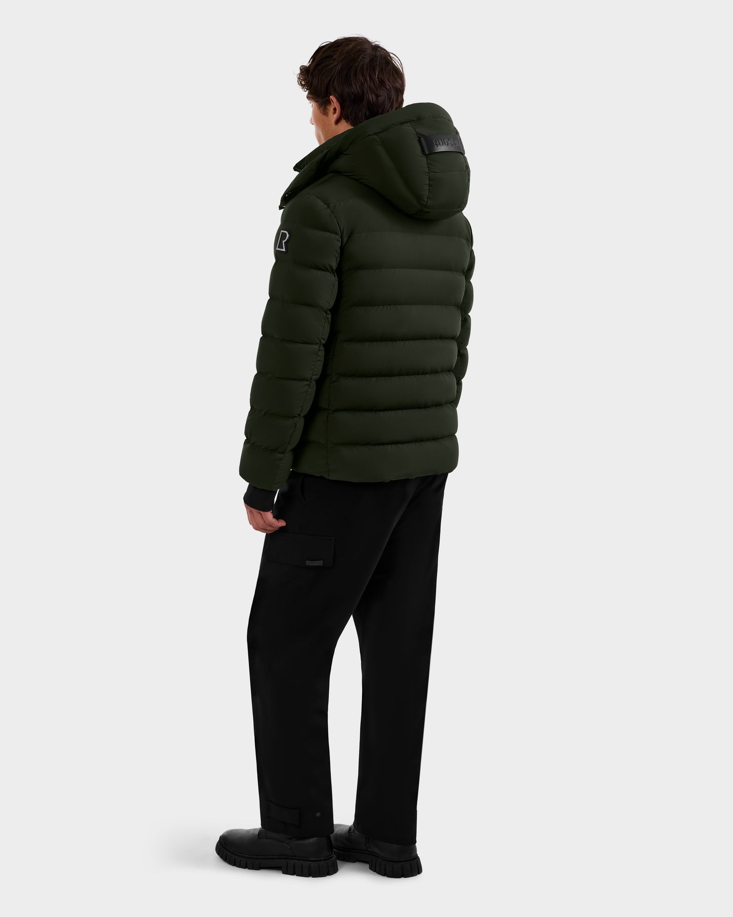 A person wearing the Rudsak NAVAN - 6125598 Dark Olive RDS-certified down quilted puffer jacket with a hood, black pants, and black shoes stands facing away from the camera against a plain light background.