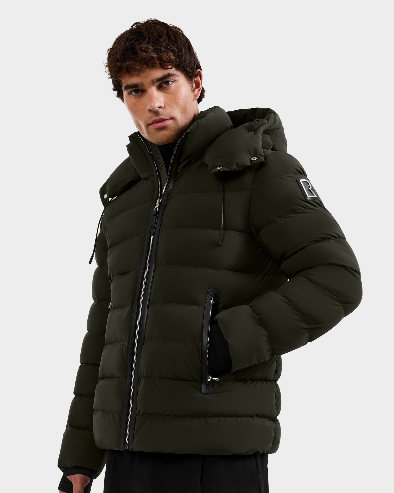 A man stands facing forward, wearing the Rudsak NAVAN - 6125598 Dark Olive men’s down puffer with a hood. Featuring a sleeve patch and front zipper, the light quilted jacket is shown against a plain, light-colored background.