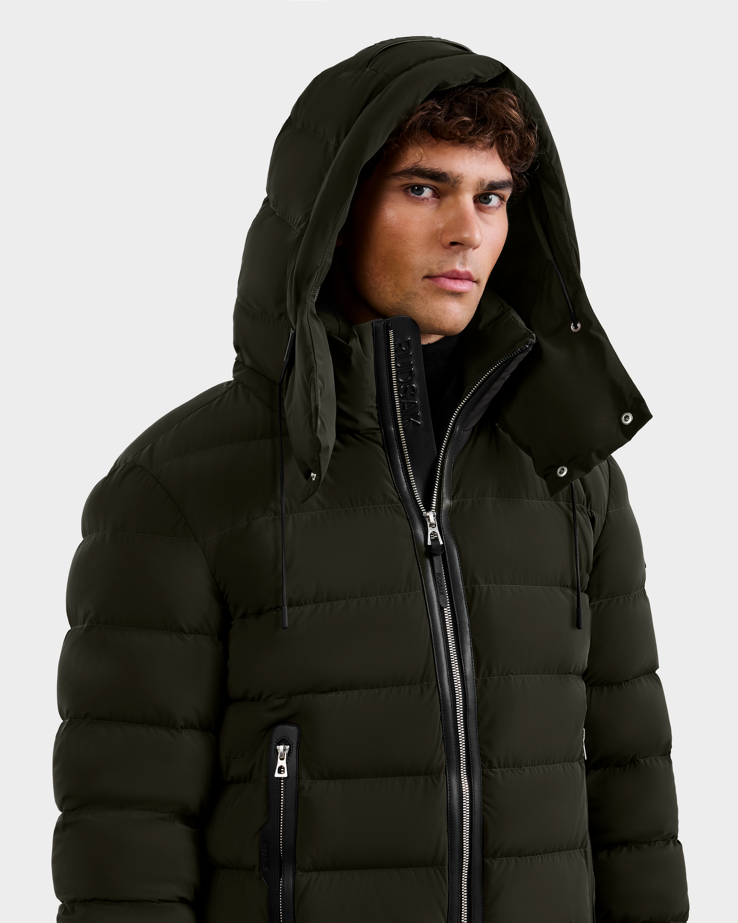 A young man wears the Rudsak NAVAN - 6125598 Dark Olive men's hooded down puffer with visible zippers and horizontal quilting. The RDS-certified jacket stands out against a plain, light background as he gazes slightly to the side.