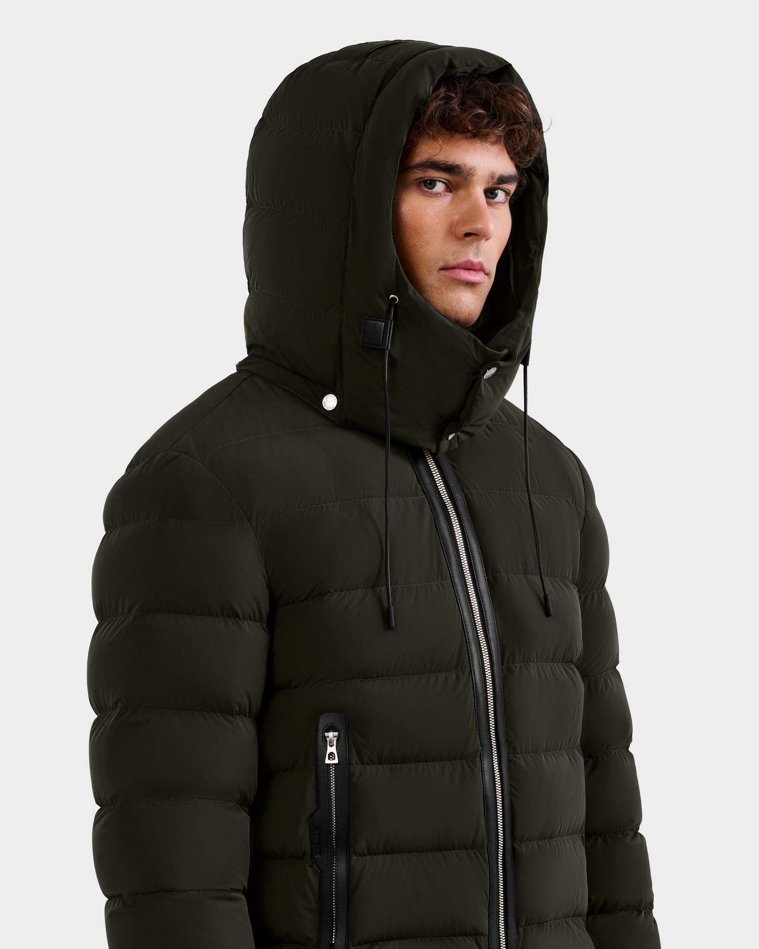 A young man wears the NAVAN - 6125598 Dark Olive down puffer by Rudsak, featuring zippered pockets, black hood drawstrings, and a hood, against a plain light gray background, looking slightly to the side.