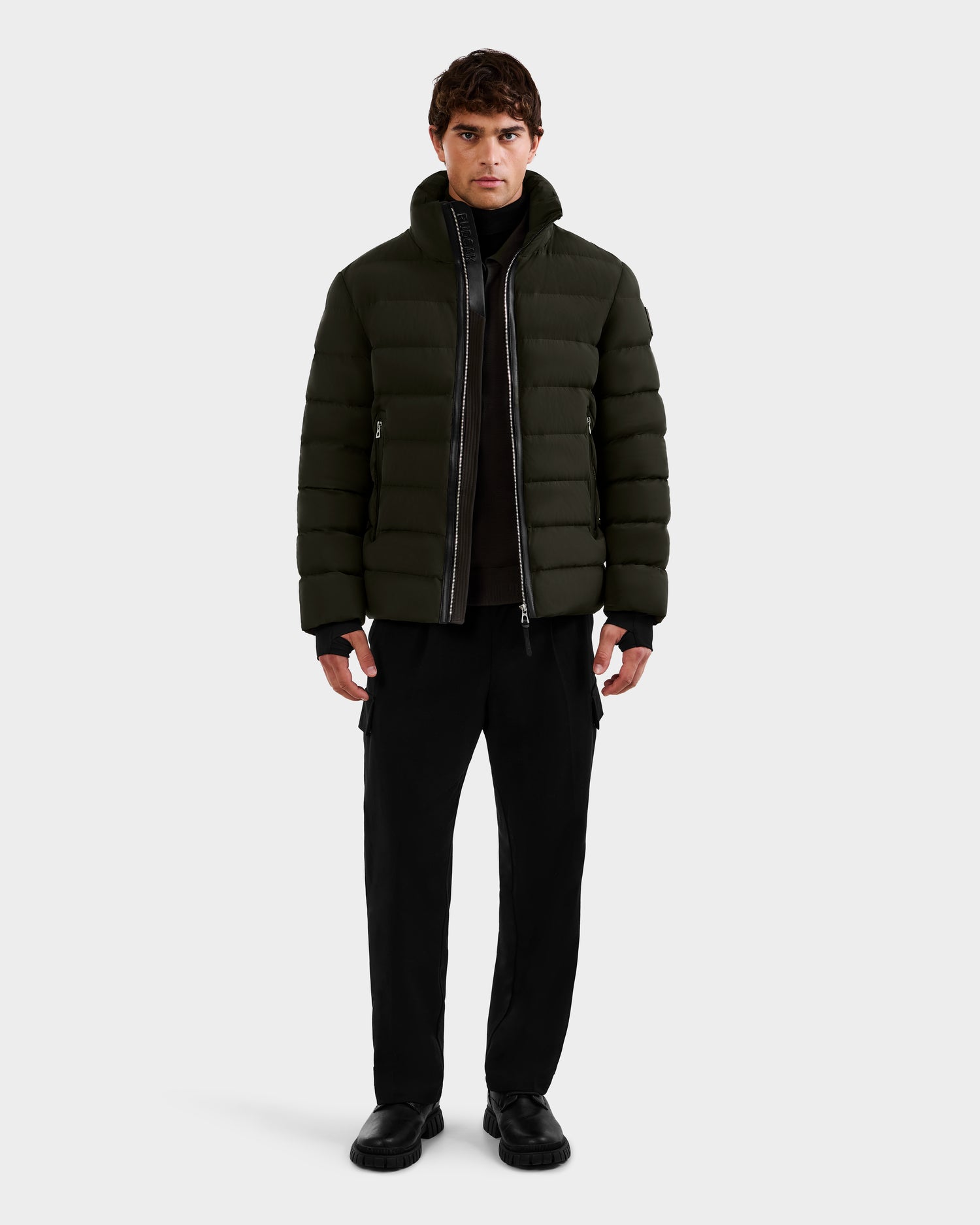 A man faces forward wearing the Rudsak NAVAN - 6125598 Dark Olive quilted puffer jacket over a black top and pants with black shoes, set against a plain light background.
