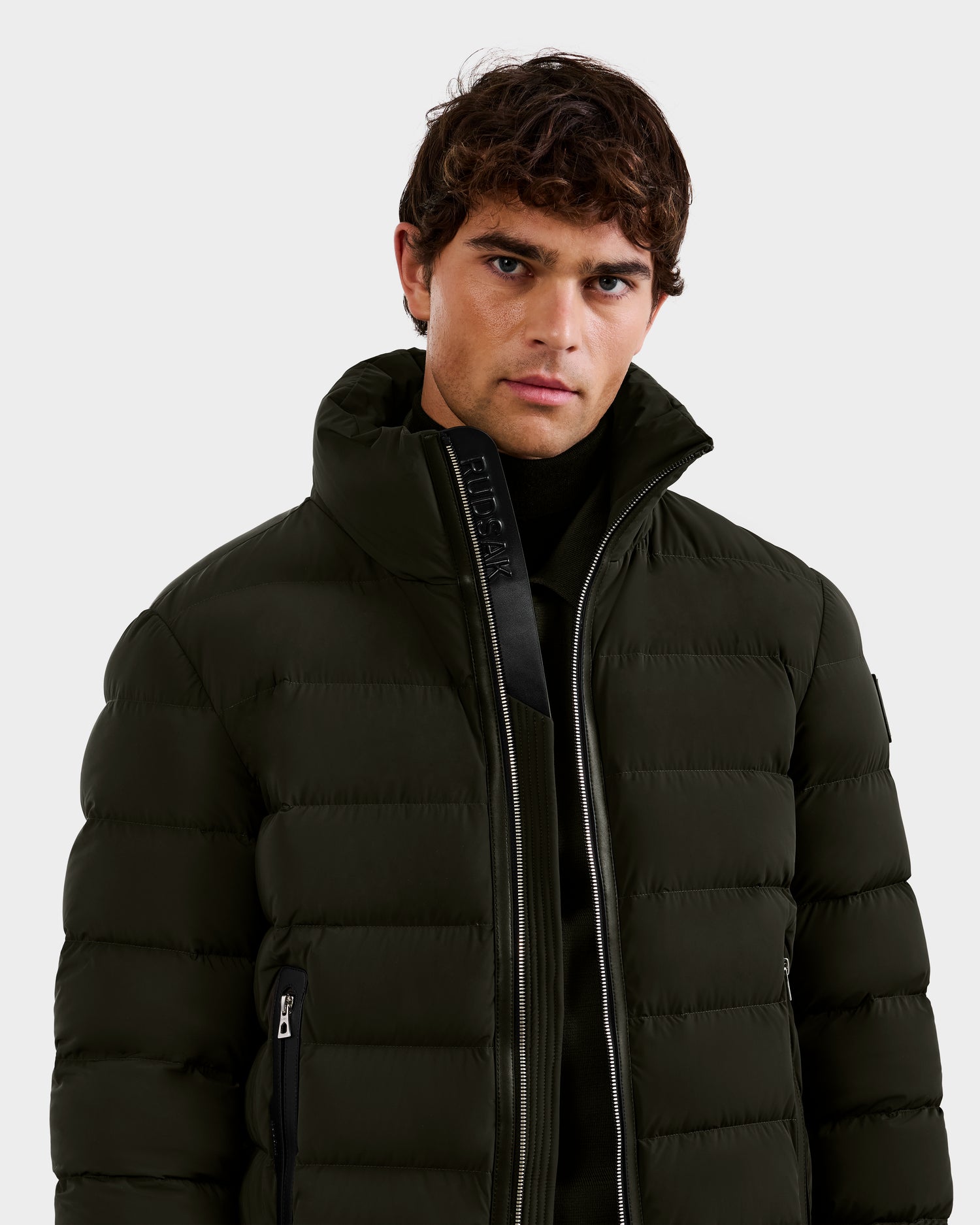 A man with short, curly brown hair wears a Rudsak NAVAN - 6125598 Dark Olive men's down puffer jacket over a black top, standing against a plain light gray background.
