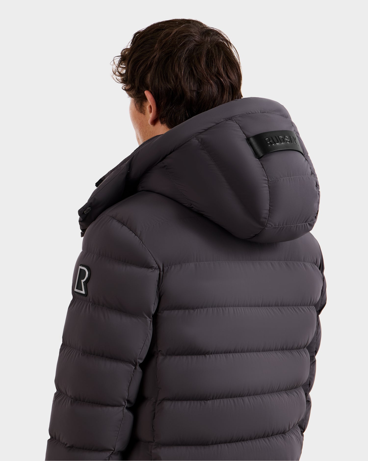 A man with short brown hair is shown from the back wearing the Rudsak NAVAN - 6125598 Grey men's quilted down puffer jacket, which is wind and water-repellent, with a sleeve patch and black label on the hood visible.