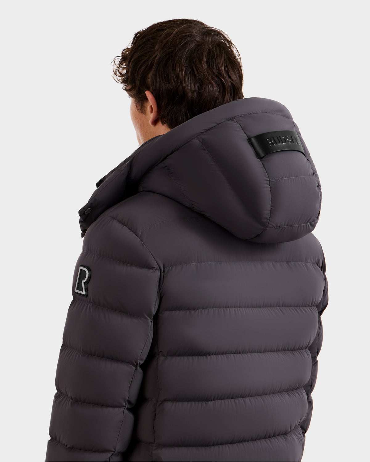 A man with short brown hair is shown from the back wearing the Rudsak NAVAN - 6125598 Grey men's quilted down puffer jacket, which is wind and water-repellent, with a sleeve patch and black label on the hood visible.