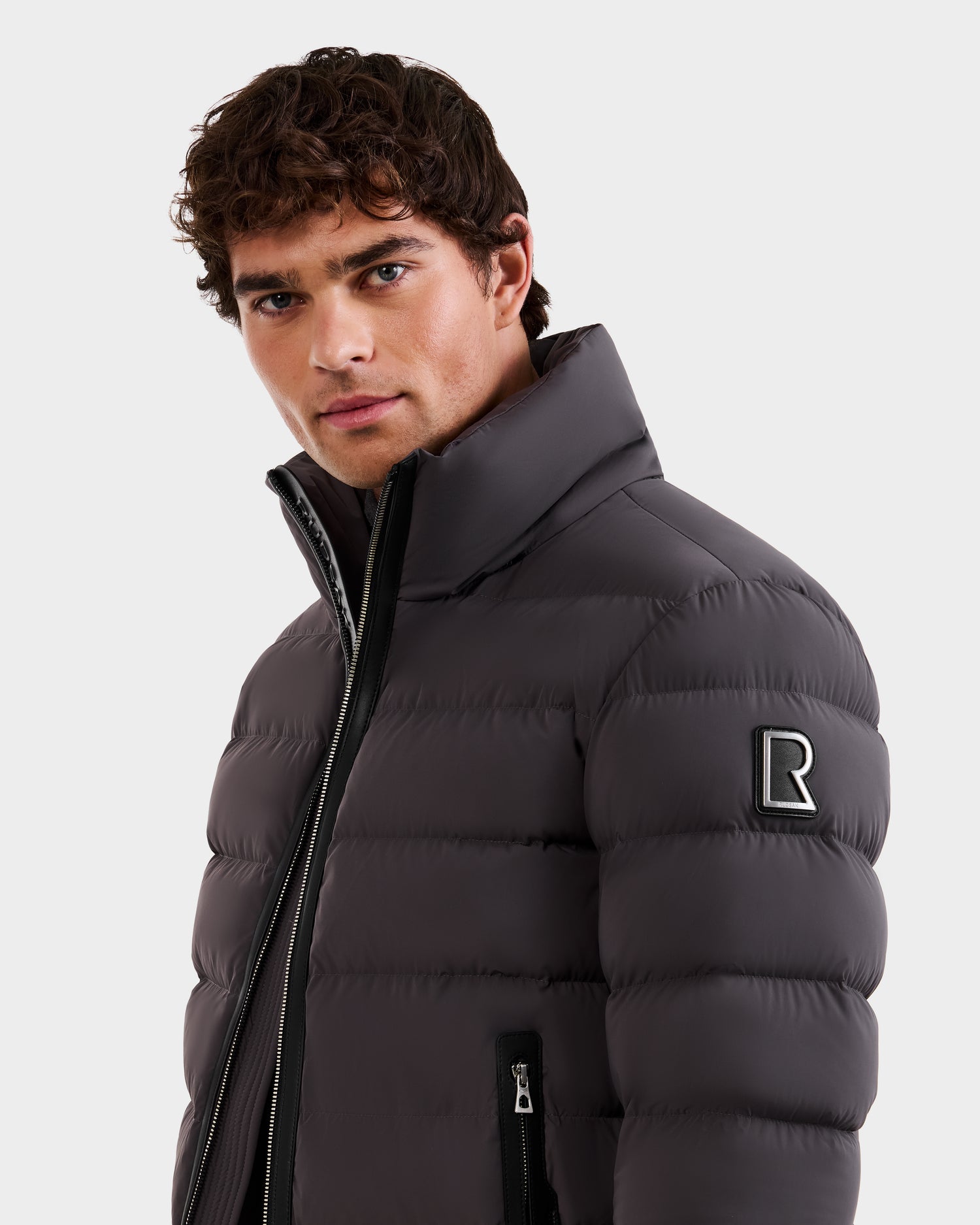 A man with curly hair wears the Rudsak NAVAN - 6125598 Grey men's quilted down puffer with a high collar and an R logo patch on the sleeve, looking slightly to the side against a plain, light background.