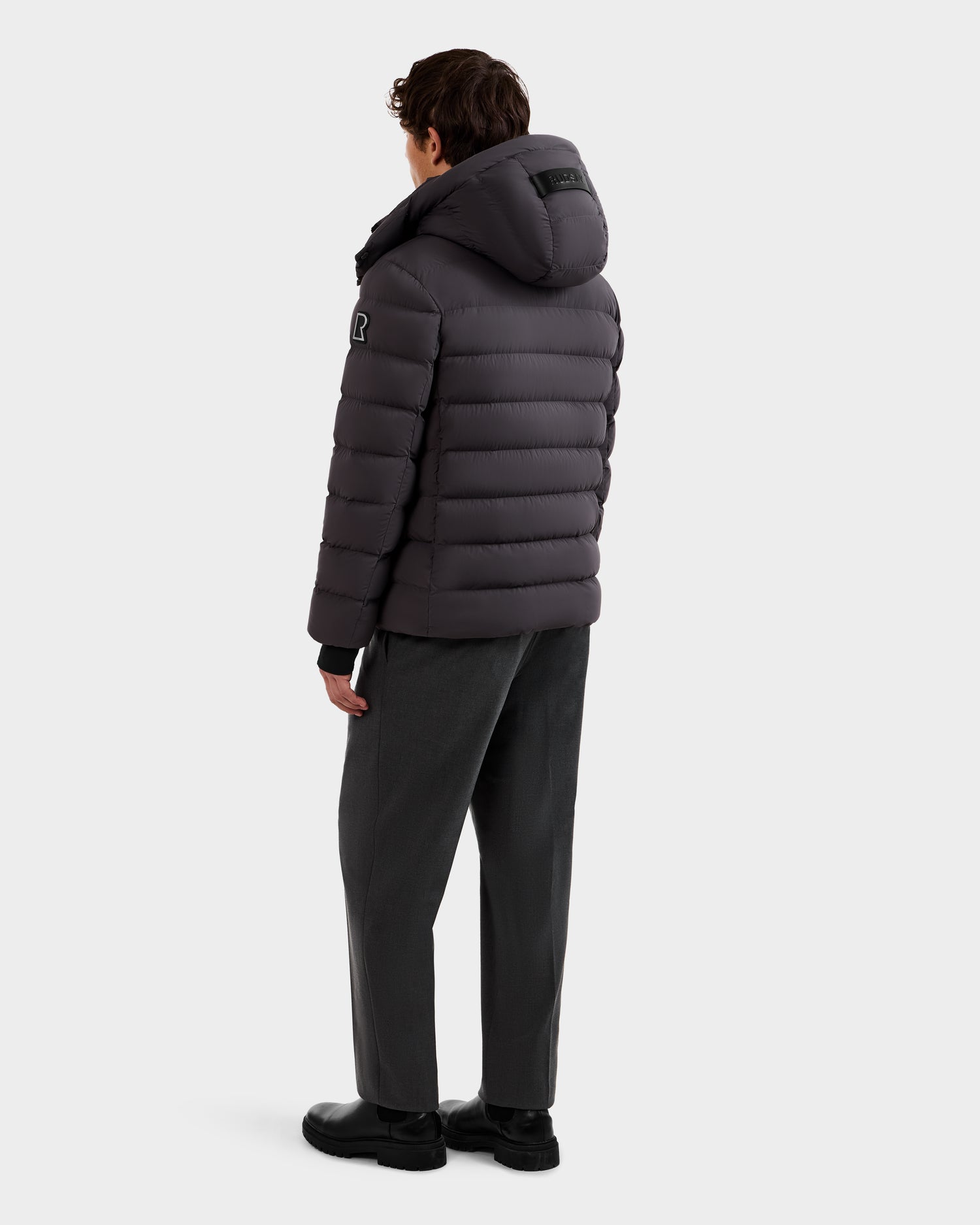 A person stands facing away in the Rudsak NAVAN - 6125598 Grey men's quilted down puffer jacket with hood, gray pants, and black shoes. The wind- and water-repellent jacket is perfect for chilly days, set against a plain light background.