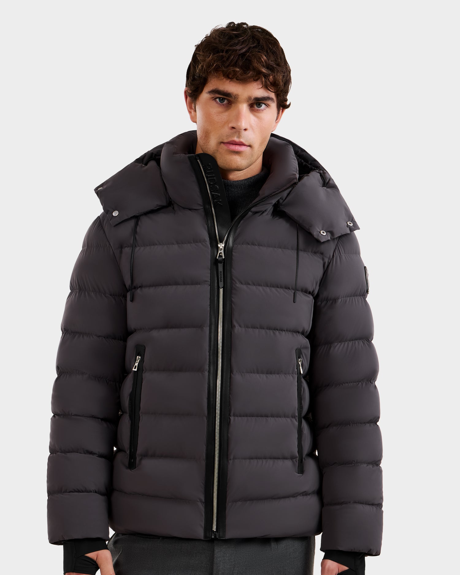 A young man with short, curly brown hair wears Rudsak’s NAVAN - 6125598 Grey men’s quilted down puffer jacket, featuring a high collar, front zipper, zippered pockets, and RDS-certified down for wind and water-repellent protection.