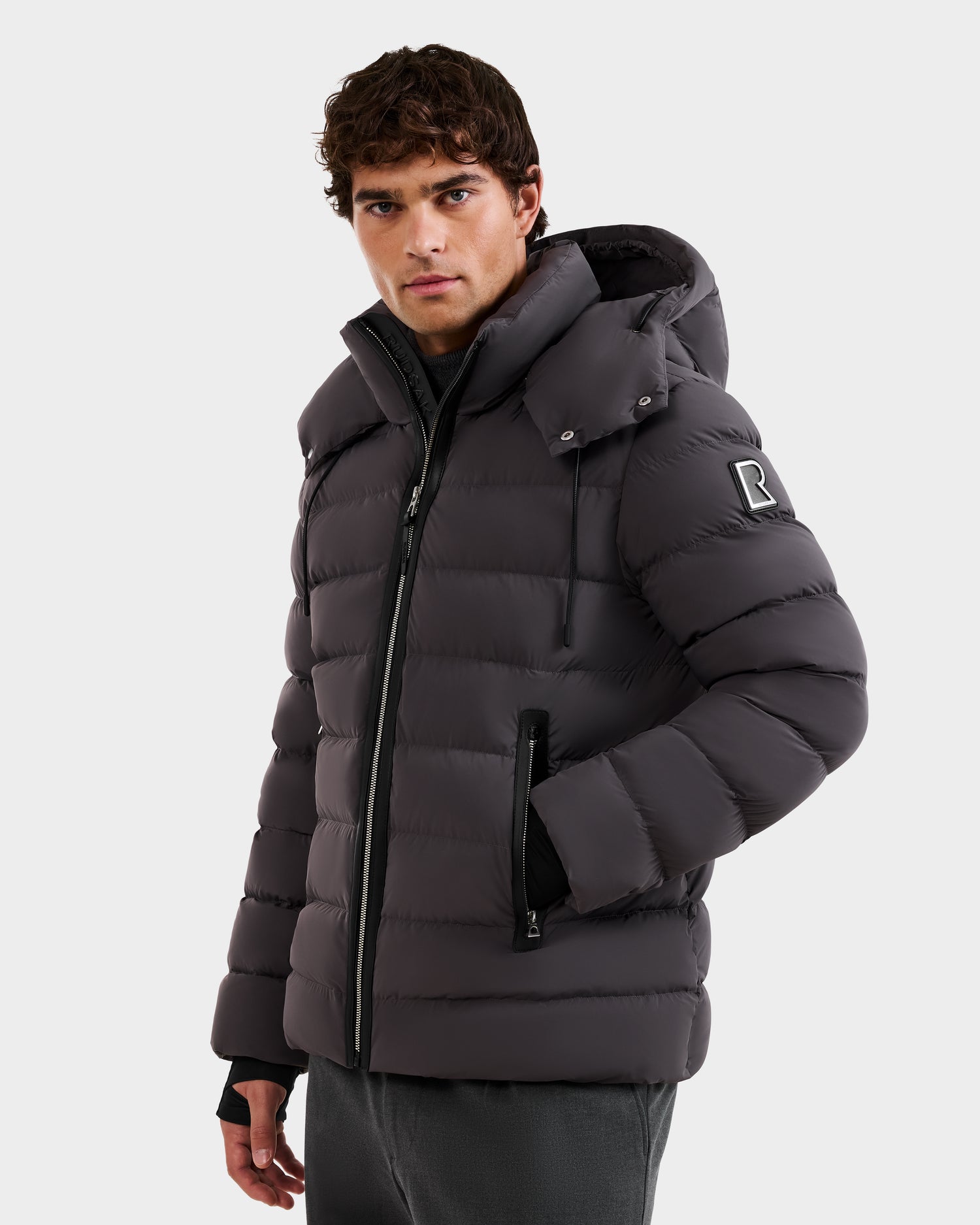 A man wears the Rudsak NAVAN - 6125598 Grey men's quilted down puffer jacket with a hood, water-repellent fabric, zip closure, front pockets, and an R patch on the left sleeve, standing against a plain white background.