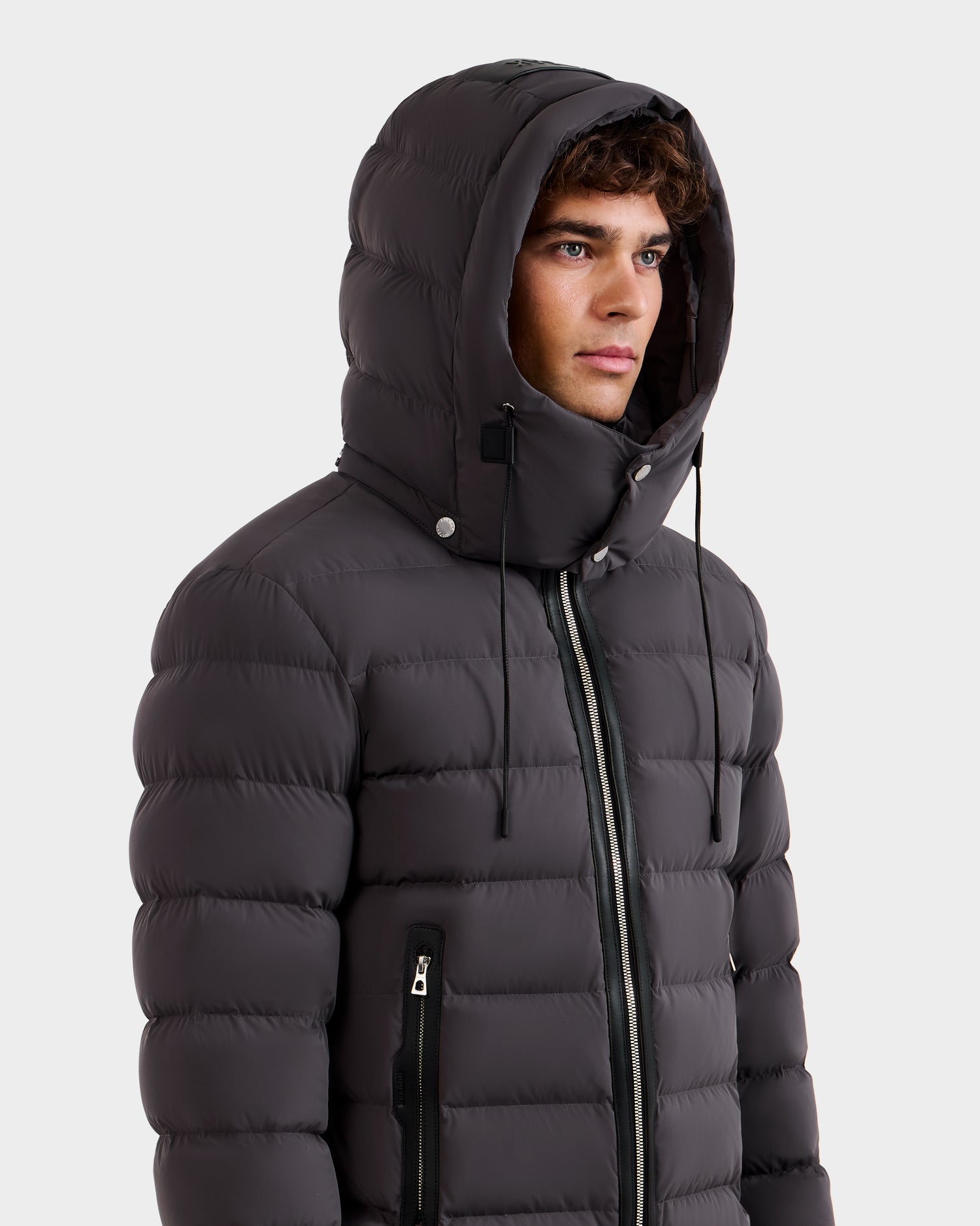 A young man wearing the Rudsak NAVAN - 6125598 Grey men's quilted down puffer with hood, made from RDS-certified down, featuring a zipper, zippered pockets, and drawstrings, stands against a plain, light background.