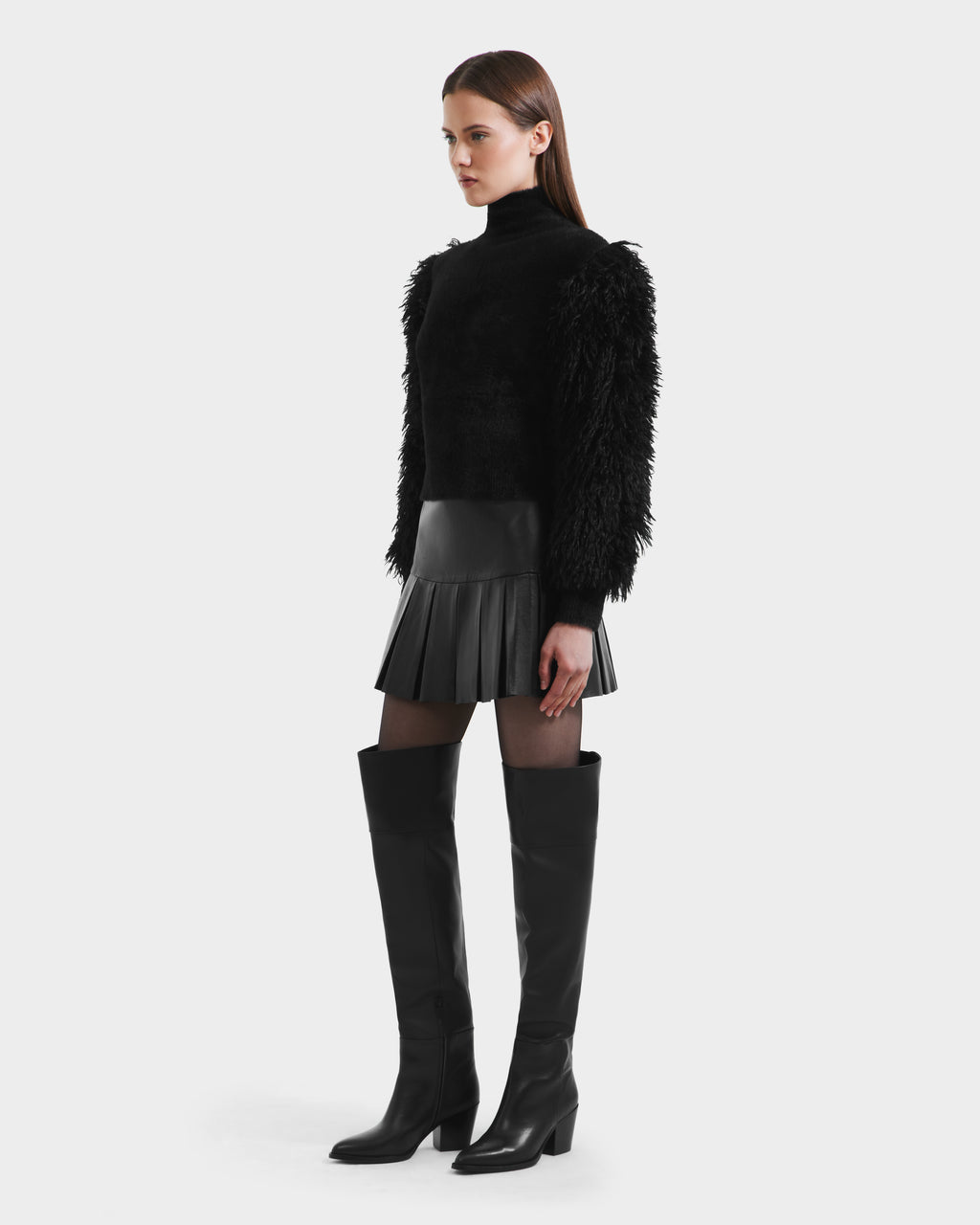 A woman stands in profile wearing the Rudsak NOEL - 8525571 Black mock-neck sweater, paired with a black pleated mini skirt, sheer tights, and heeled knee-high boots against a plain white background for a chic winter look.