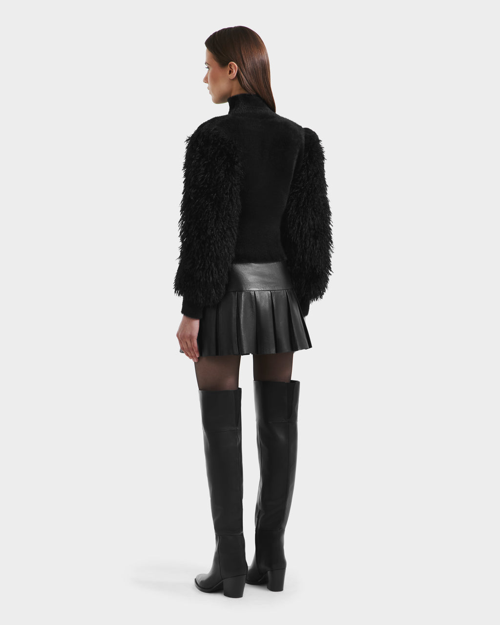 A woman faces away, wearing the NOEL - 8525571 Black jacket by Rudsak with faux fur sleeves, a pleated leather mini skirt, sheer black tights, and knee-high boots against a plain white background.