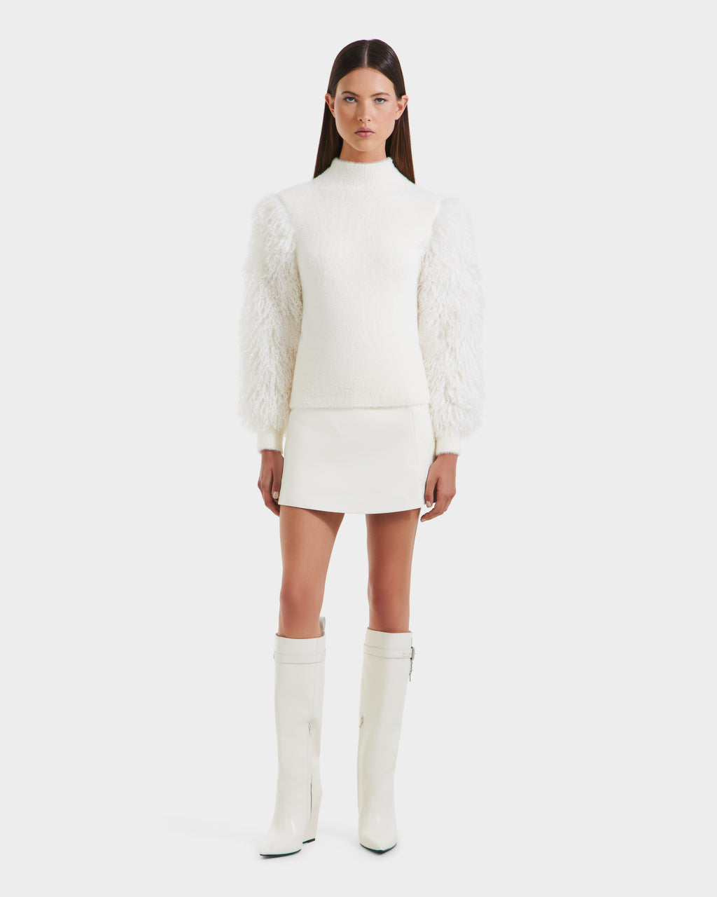 A woman with long brown hair models Rudsak’s NOEL - 8525571 Vanilla: a chic women’s mock-neck sweater with voluminous faux fur sleeves, styled with a white mini skirt and knee-high boots, against a plain white background.