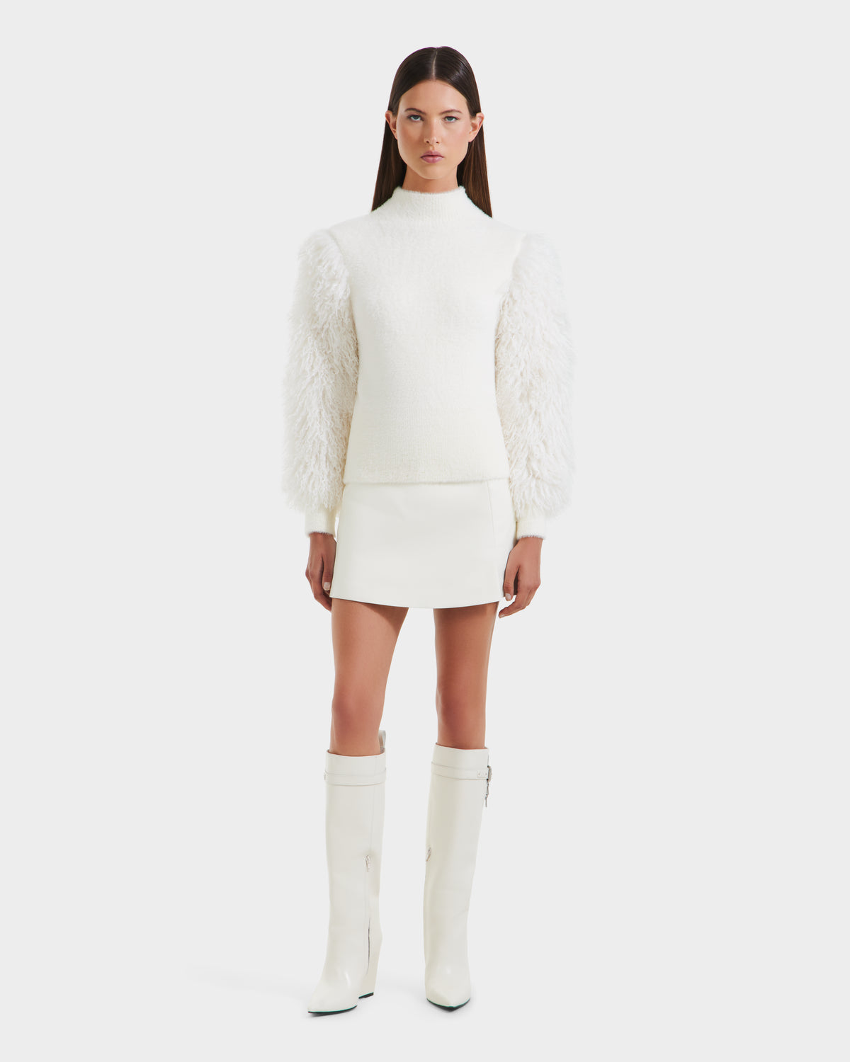 A woman with long brown hair models Rudsak’s NOEL - 8525571 Vanilla: a chic women’s mock-neck sweater with voluminous faux fur sleeves, styled with a white mini skirt and knee-high boots, against a plain white background.
