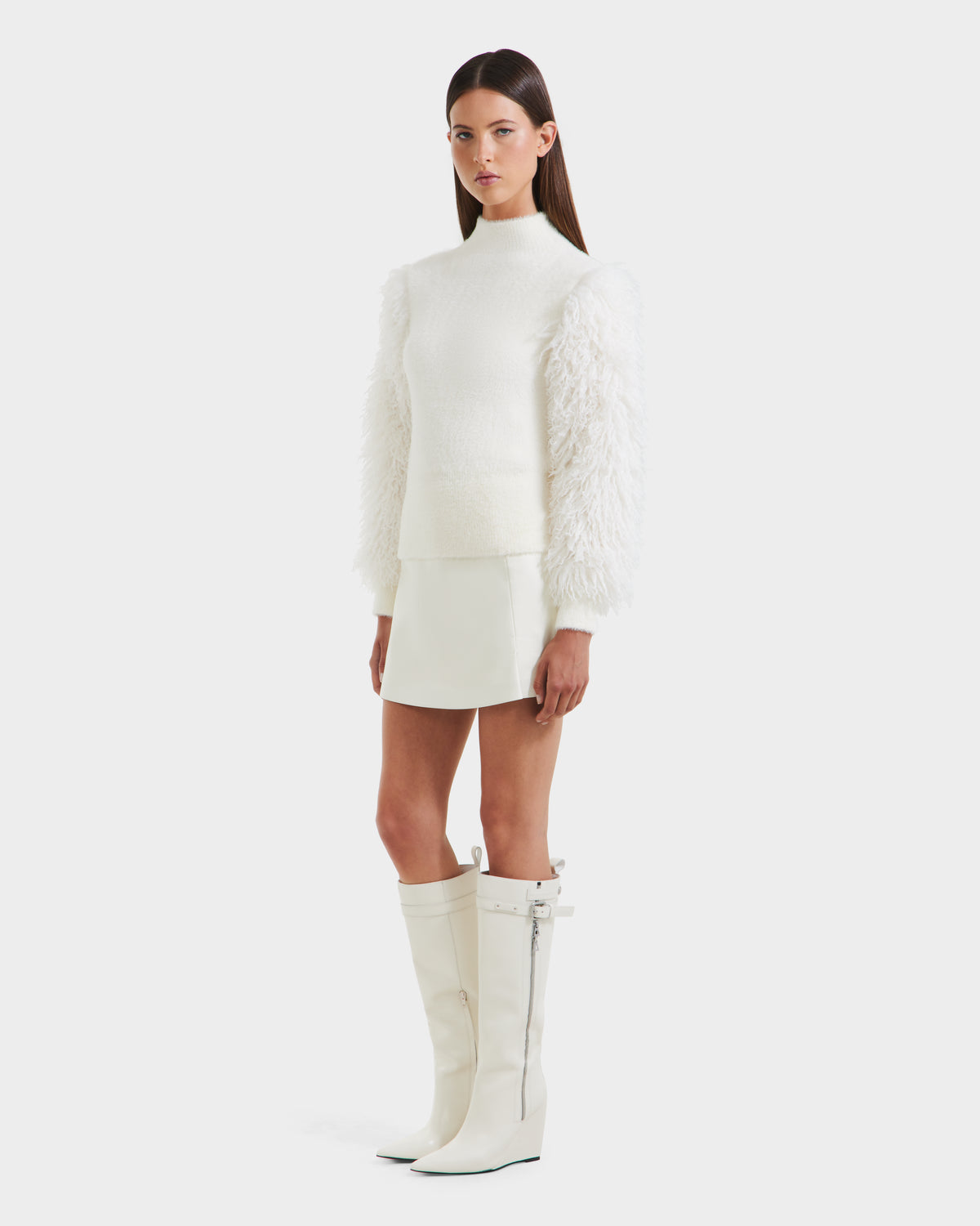 A woman with long straight hair and a neutral expression stands against a plain background wearing the Rudsak NOEL - 8525571 Vanilla mock-neck sweater with faux fur sleeves, white mini skirt, and tall white wedge boots for chic winter style.