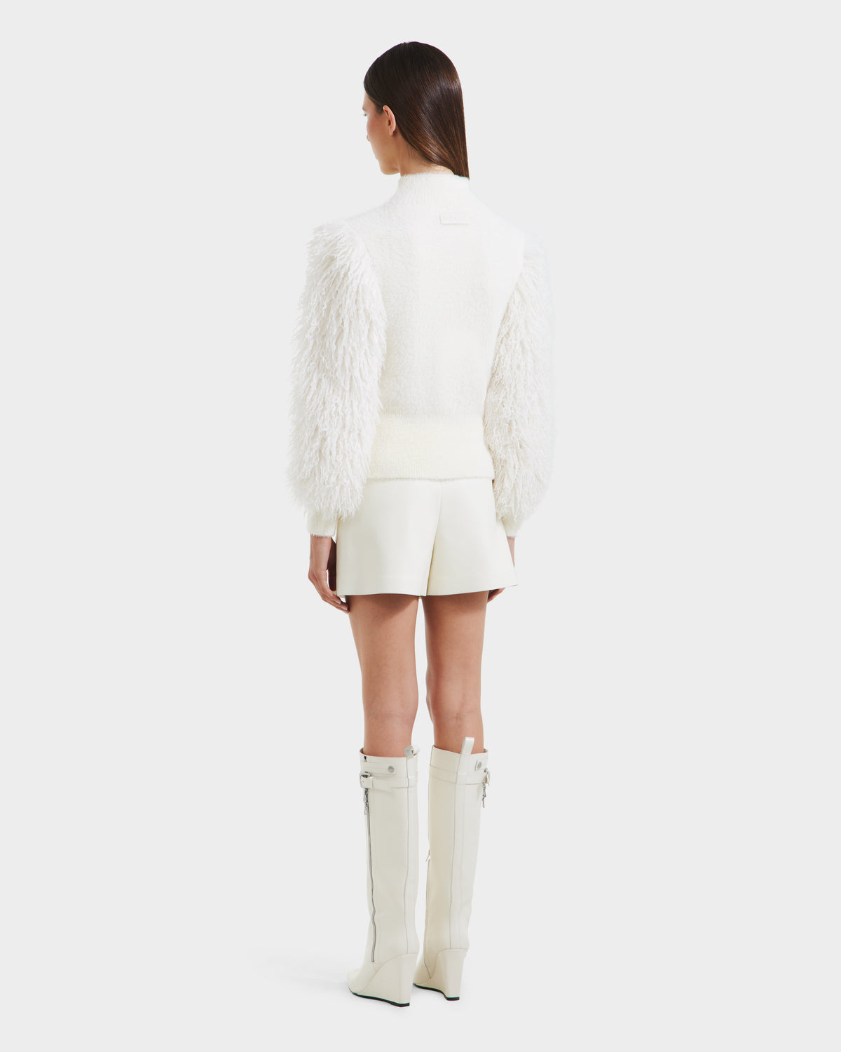 A woman faces away, wearing the Rudsak NOEL - 8525571 Vanilla faux fur mock-neck sweater, a short white skirt, and zippered knee-high white boots, highlighting chic winter style against a plain white background.
