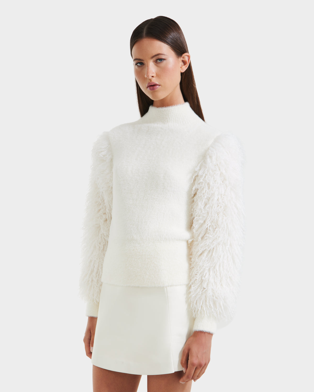 A woman wears the Rudsak NOEL - 8525571 Vanilla sweater with faux fur sleeves and a white mini skirt, standing against a plain light background.