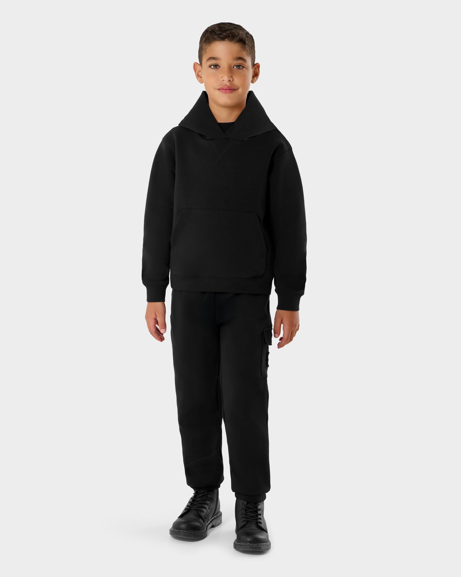 A young boy faces forward against a plain light gray background, dressed in Rudsak's NOOR - 1525004 Black hoodie, paired with black pants and black boots.