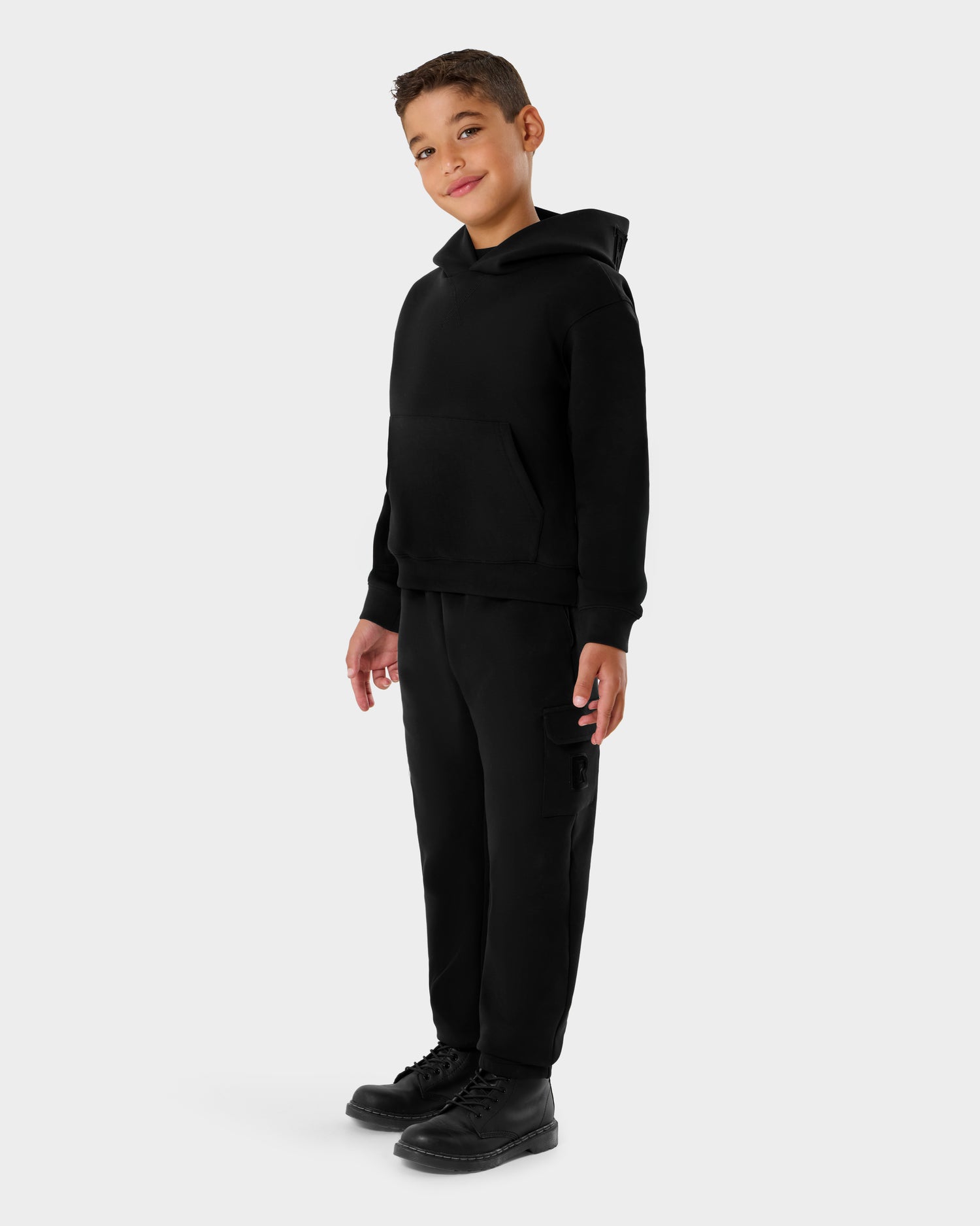 A young boy smiles, wearing the Rudsak NOOR - 1525004 Black hoodie with matching black cargo pants and boots, set against a plain light gray background.