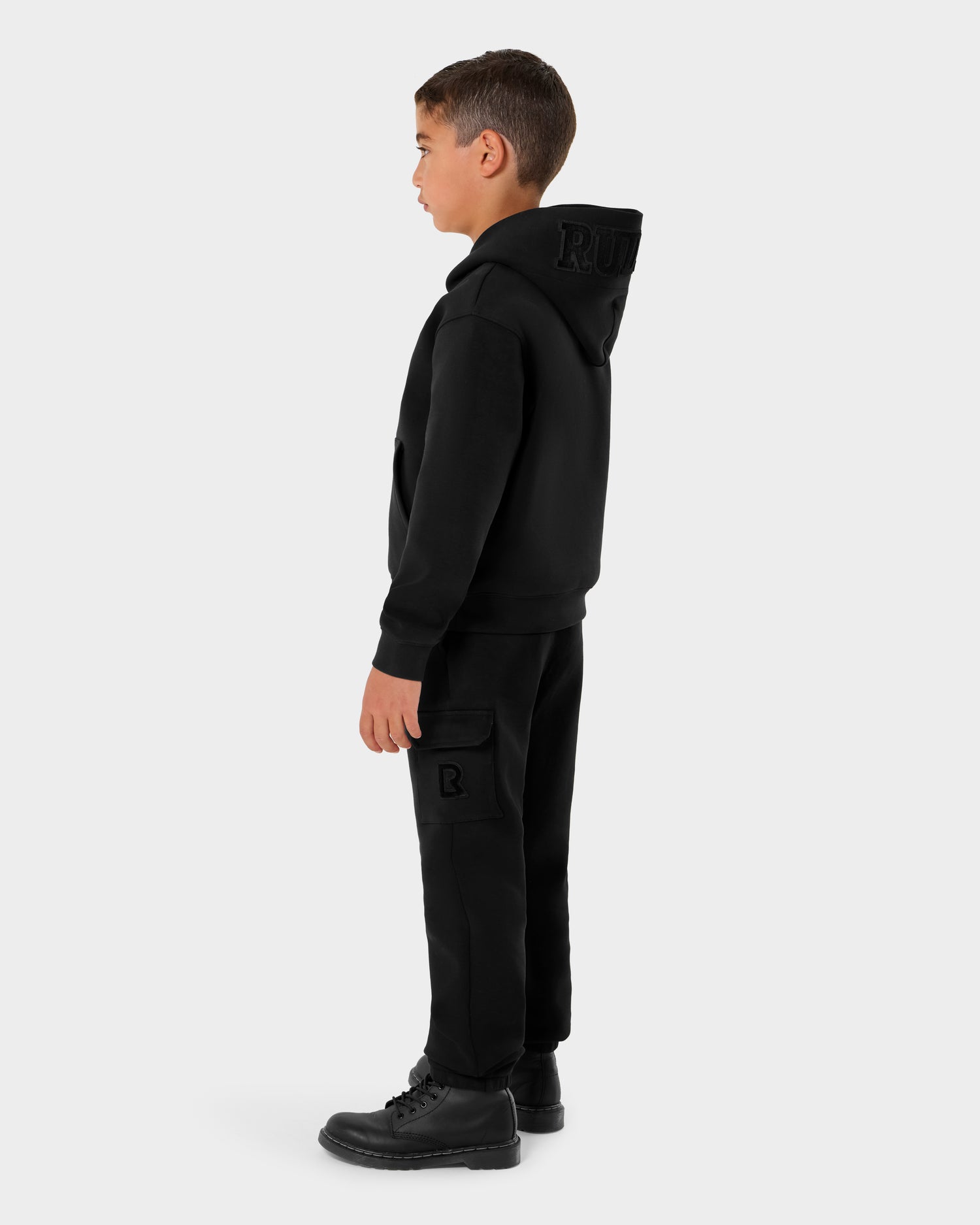 A young boy stands in profile wearing the Rudsak NOOR - 1525004 Black hoodie and cargo pants with boots. The hoodie features a raised R on the back of the hood, while the pants have a flap pocket with an R patch. Background: plain light gray.