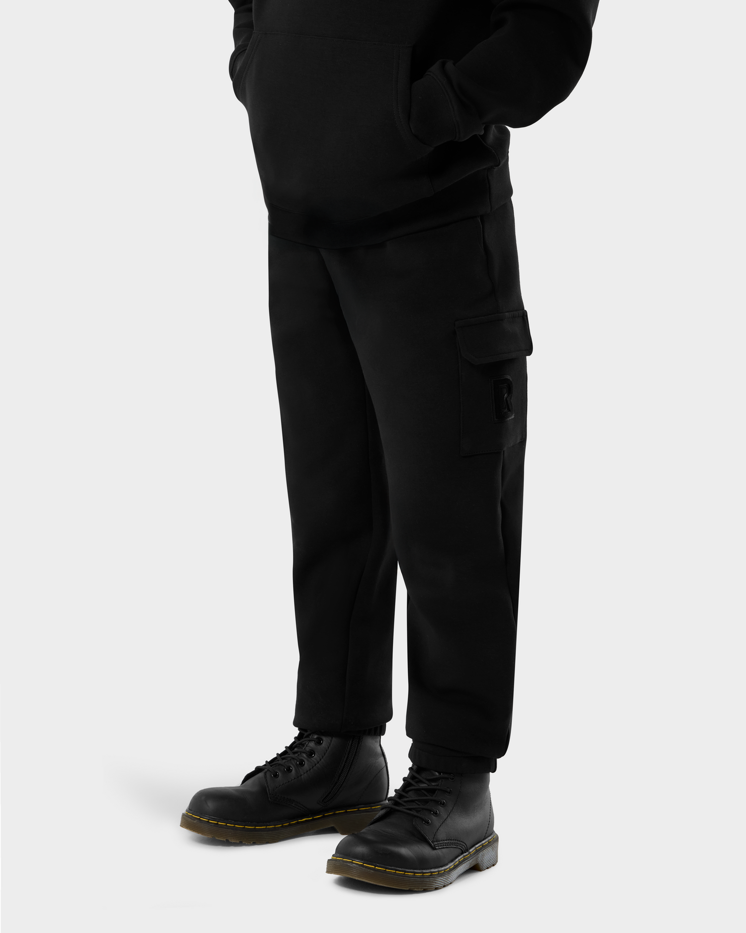 A person wears Rudsak's NOOR - 1525004 Black cargo pants with side pockets and black lace-up boots, standing against a plain light gray background. Their hands are in the front pocket of a black hoodie, face partially out of frame.