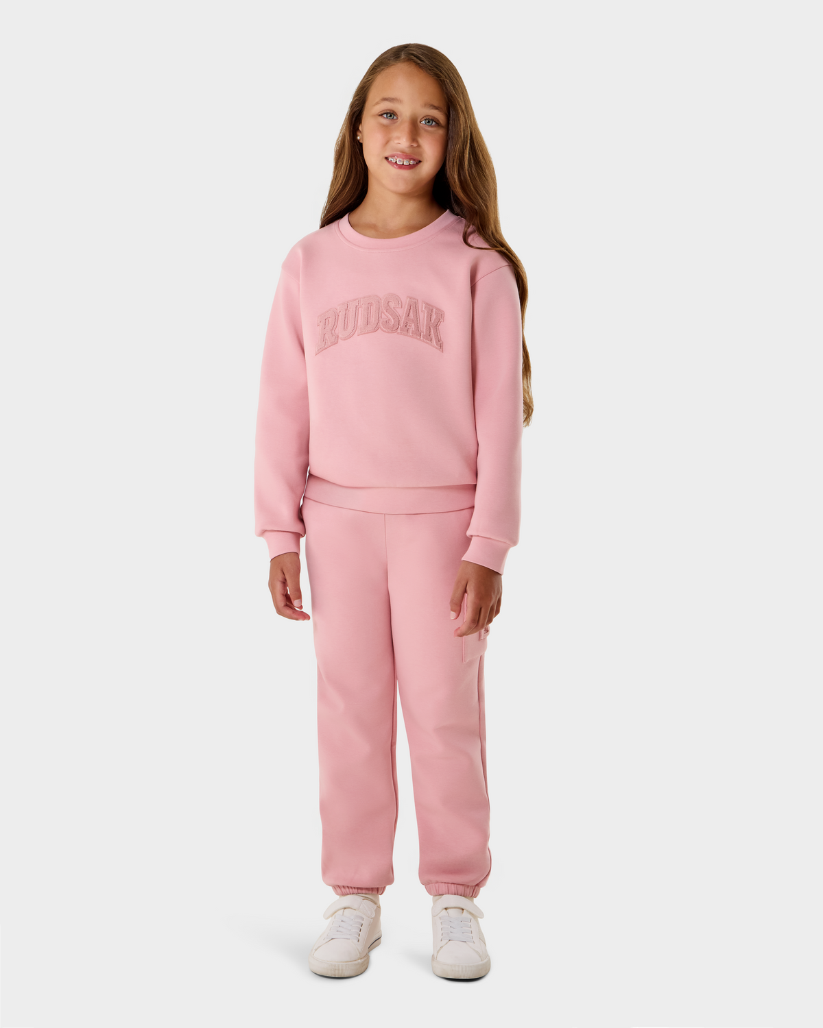A young girl with long brown hair smiles in a light pink Rudsak NOOR - 1525004 sweatshirt and sweatpants, paired with white sneakers, standing against a plain white background.