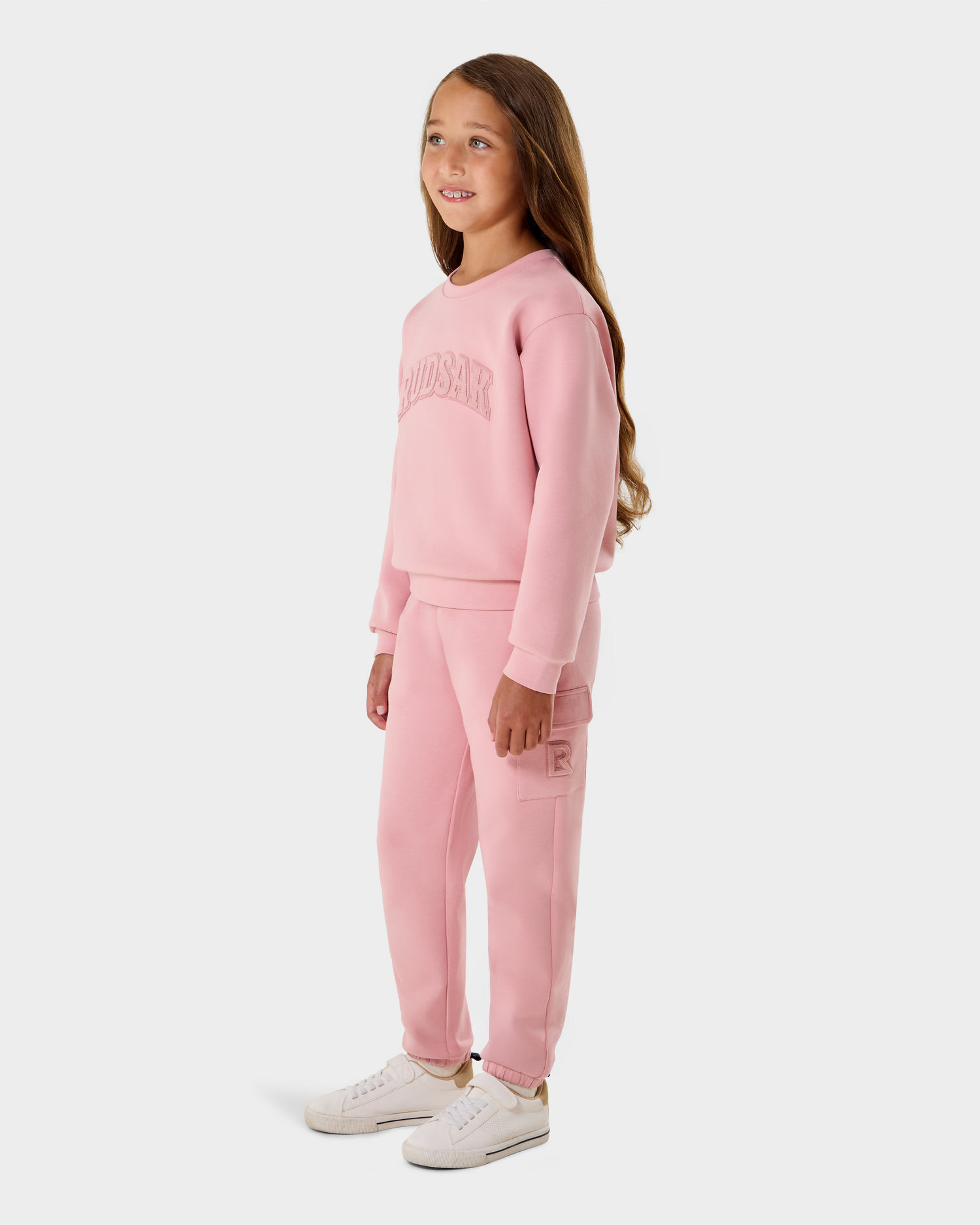 A young girl with long brown hair wears the Rudsak NOOR - 1525004 Light Pink sweatshirt and matching sweatpants with white sneakers, standing and smiling against a plain white background.