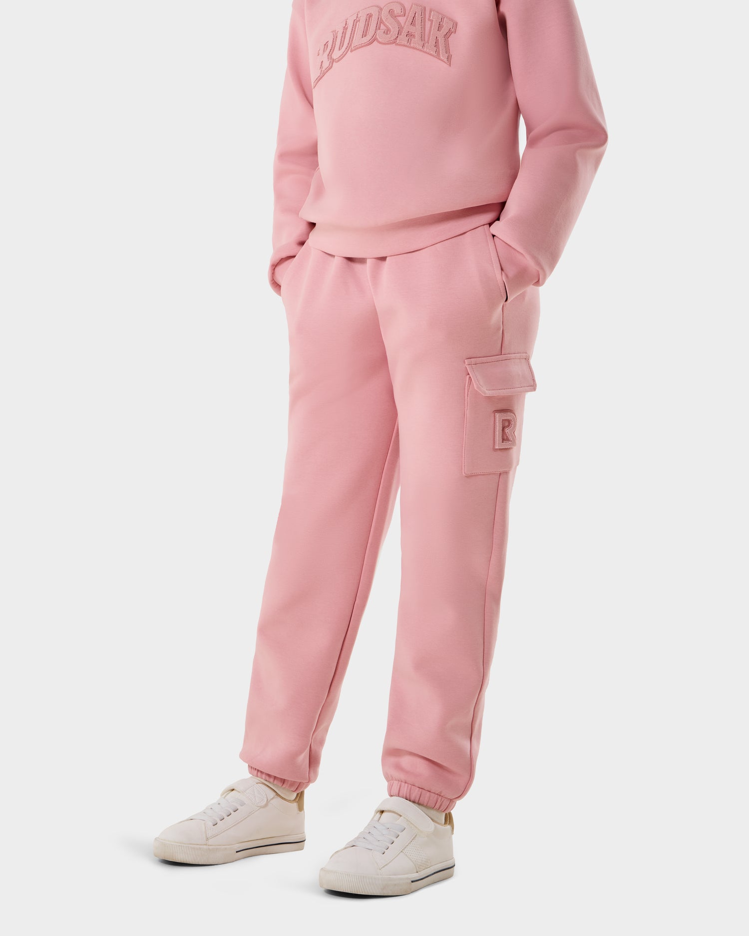 A person wears the Rudsak NOOR - 1525004 light pink sweatshirt and matching cargo jogger pants, paired with white sneakers, standing against a plain white background.
