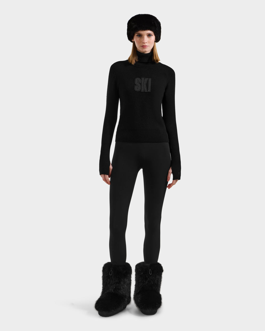 A woman faces forward, wearing the Rudsak NOVIE - 8525659 Black knit turtleneck ski graphic sweater with SKI on the front, black leggings, a black fur hat, and black fur boots against a plain white background.