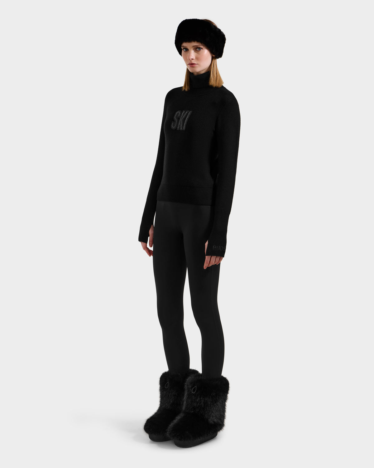 A woman stands against a white background in an all-black look, featuring the Rudsak NOVIE - 8525659 Black women's knit turtleneck ski graphic sweater with "SKI" on the front, styled with fitted pants, a fur hat, and fur-trimmed boots.