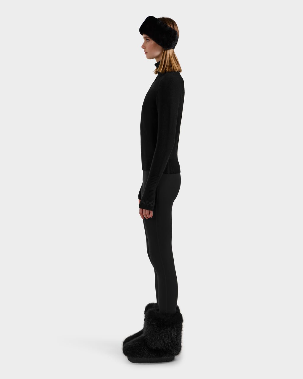 A woman in profile against a plain background wears the Rudsak NOVIE - 8525659 Black, styled with matching leggings, a fur headband, and large furry boots.