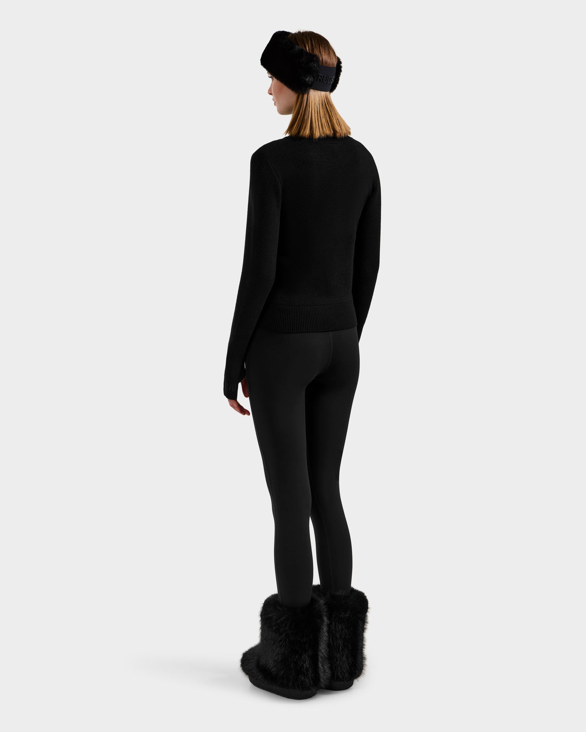 A woman faces away against a light gray background, dressed in Rudsak's NOVIE - 8525659 Black après-ski pullover with matching black leggings, furry boots, and a furry headband.