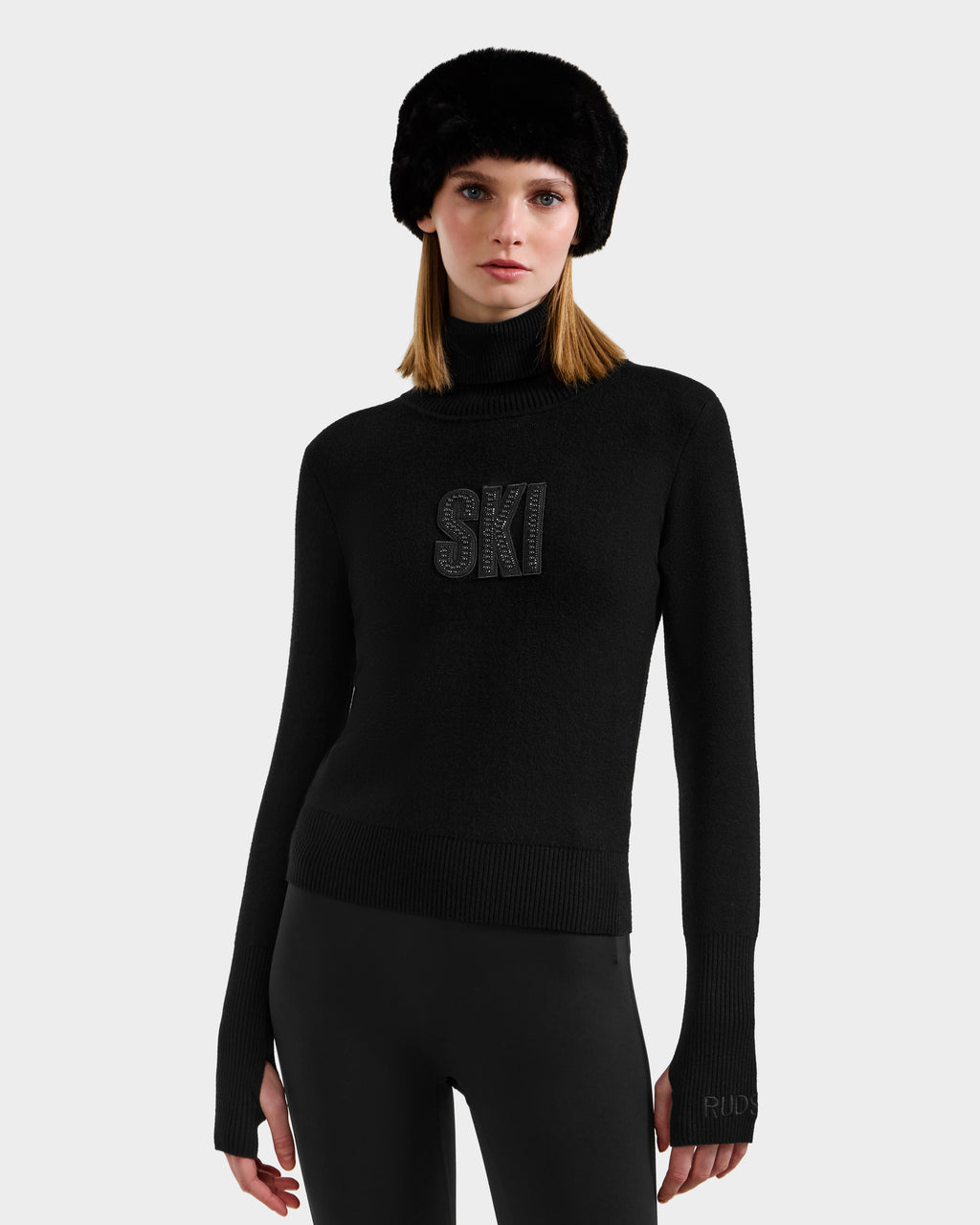 A woman stands against a light gray background wearing the Rudsak NOVIE - 8525659 Black après-ski pullover with "SKI" on the front, paired with black leggings and a black fur hat.