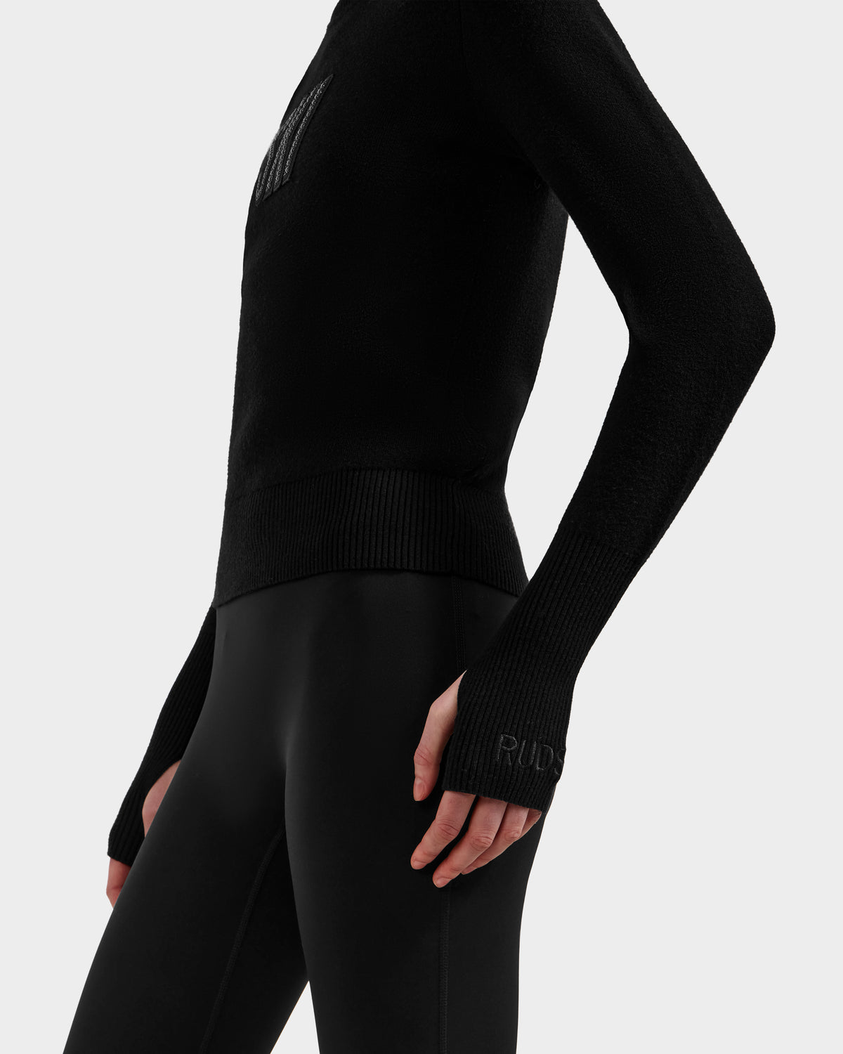 A person wears the Rudsak NOVIE - 8525659 Black women’s knit turtleneck and black leggings, cropped from shoulders to mid-thigh. One arm is bent with fingers visible, and a subtle “RUD” print is on the sleeve cuff. Background is plain white.
