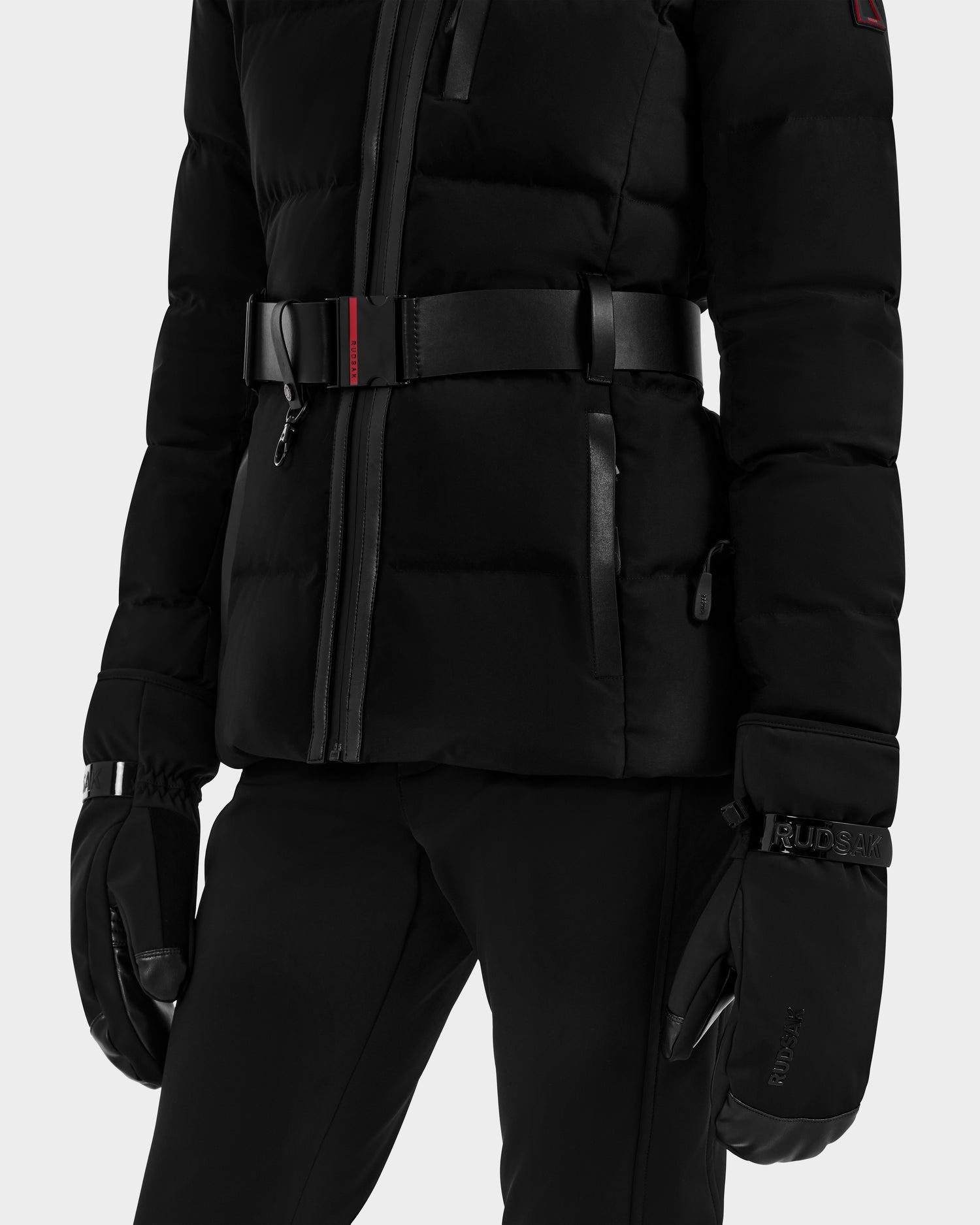 Wearing the Rudsak OLTEN - 8424513 Black puffer jacket with a zipper, red-accented belt, and zippered pockets, paired with black pants and insulated gloves—an outfit made for cold weather.
