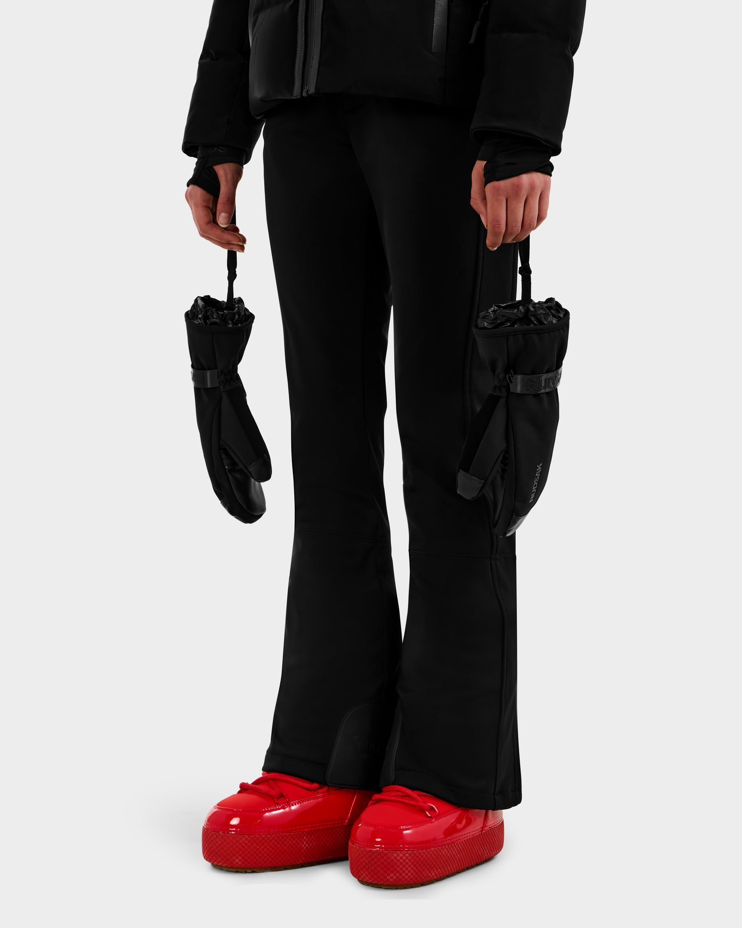 A person wearing the OLTEN - 9425565 Black pants by Rudsak stands against a plain white background, holding ski mittens by their straps with arms relaxed at their sides, paired with a black puffer jacket and bright red snow boots.