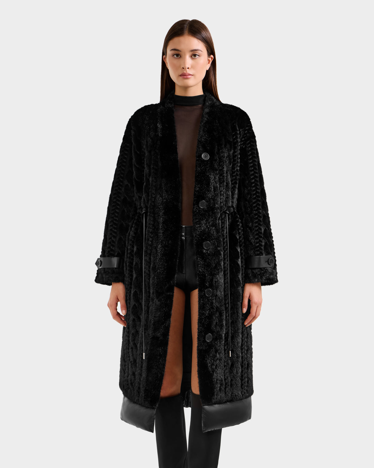 A woman with straight brown hair wears the ORIANA - 8125545 Black women’s faux fur coat by Rudsak, featuring vegan leather trims and front buttons. She pairs it with black shorts and a sheer black top against a light gray background.