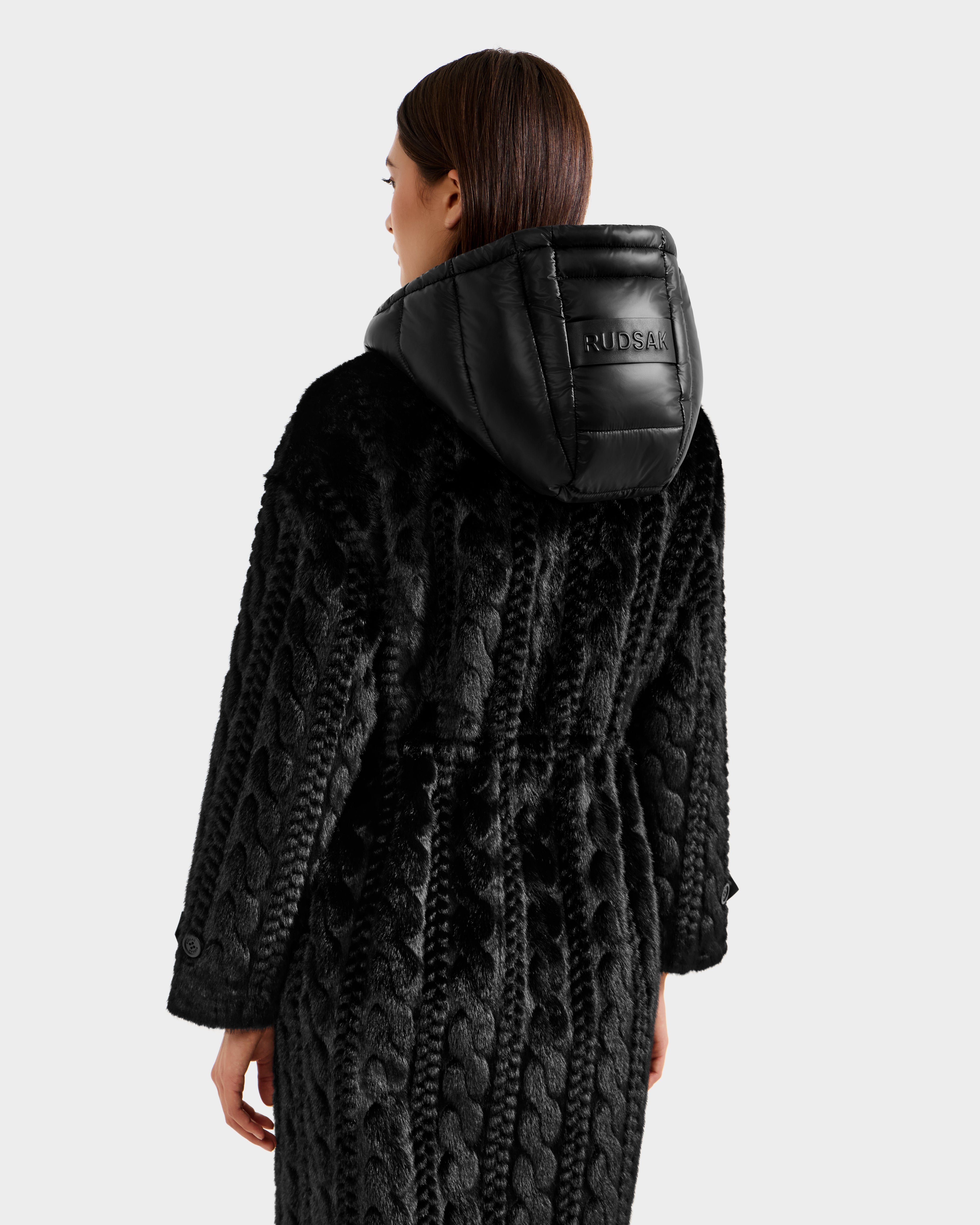 A woman with straight brown hair wears the Rudsak ORIANA - 8125545 Black coat with a quilted hood, featuring vegan leather trims on the back and hood, as she faces away.