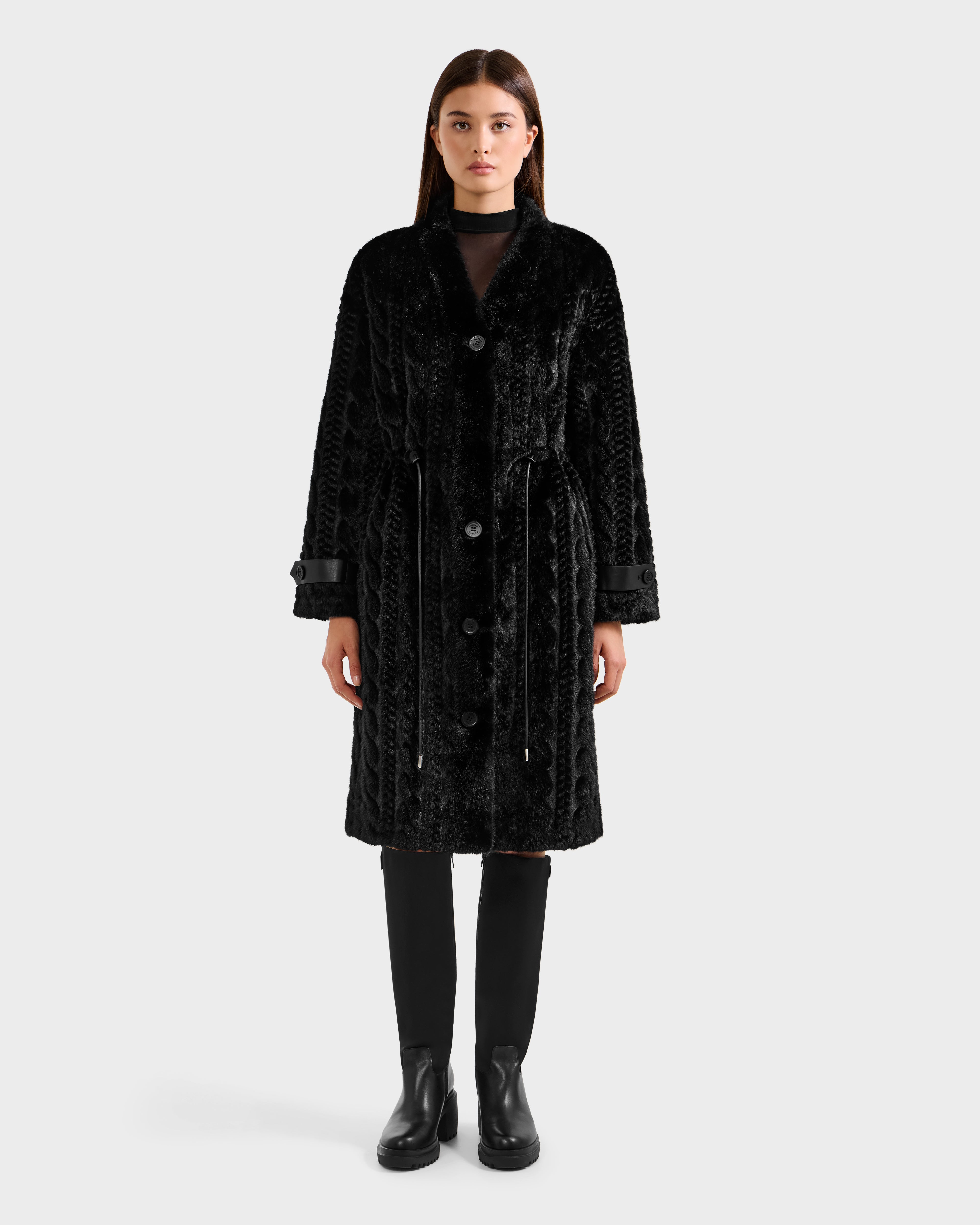 A woman faces forward against a light gray background, wearing the Rudsak ORIANA - 8125545 Black knee-length textured coat with vegan leather trim, large buttons, a belt, and black knee-high boots.