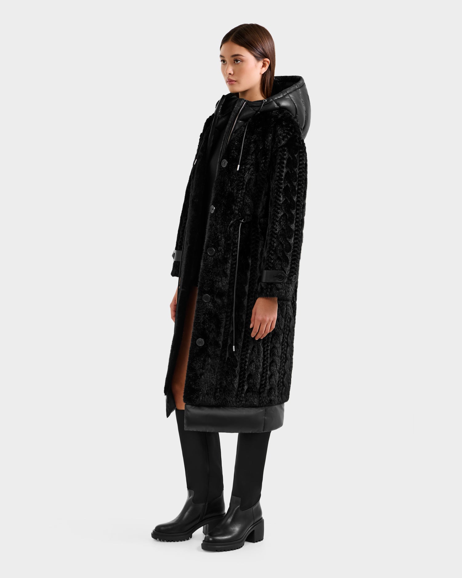 A woman wears the Rudsak ORIANA - 8125545 Black faux fur coat with a hood and vegan leather trims, paired with black boots. A brown inner layer is visible at the hem as she faces slightly right against a plain light background.