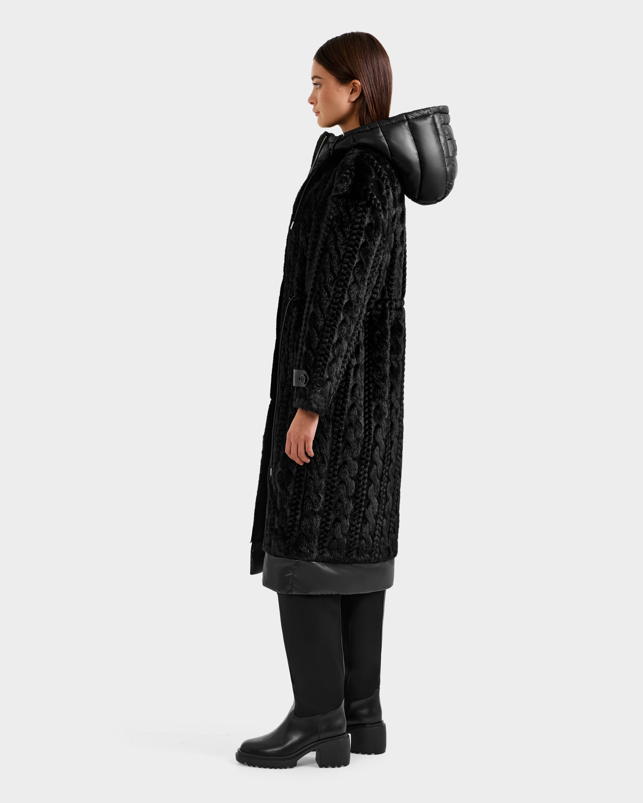 A woman in profile wears the Rudsak ORIANA - 8125545 Black textured coat with vegan leather trims, paired with black pants and boots, standing against a plain light gray background.