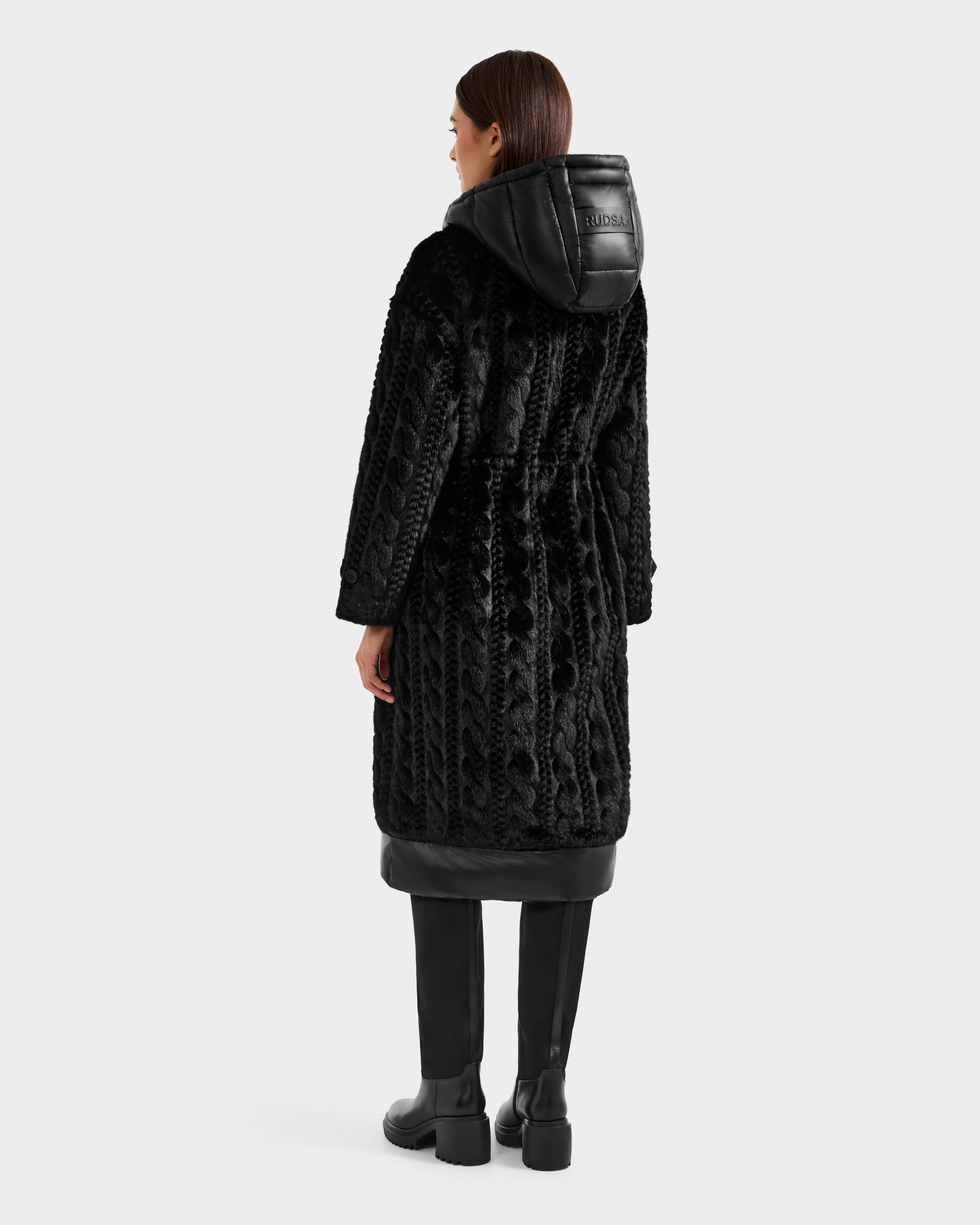 A woman faces away, wearing the Rudsak ORIANA - 8125545 Black women’s faux fur coat with a hood, vegan leather trims, and lettering. She pairs it with knee-high black boots against a plain white background.