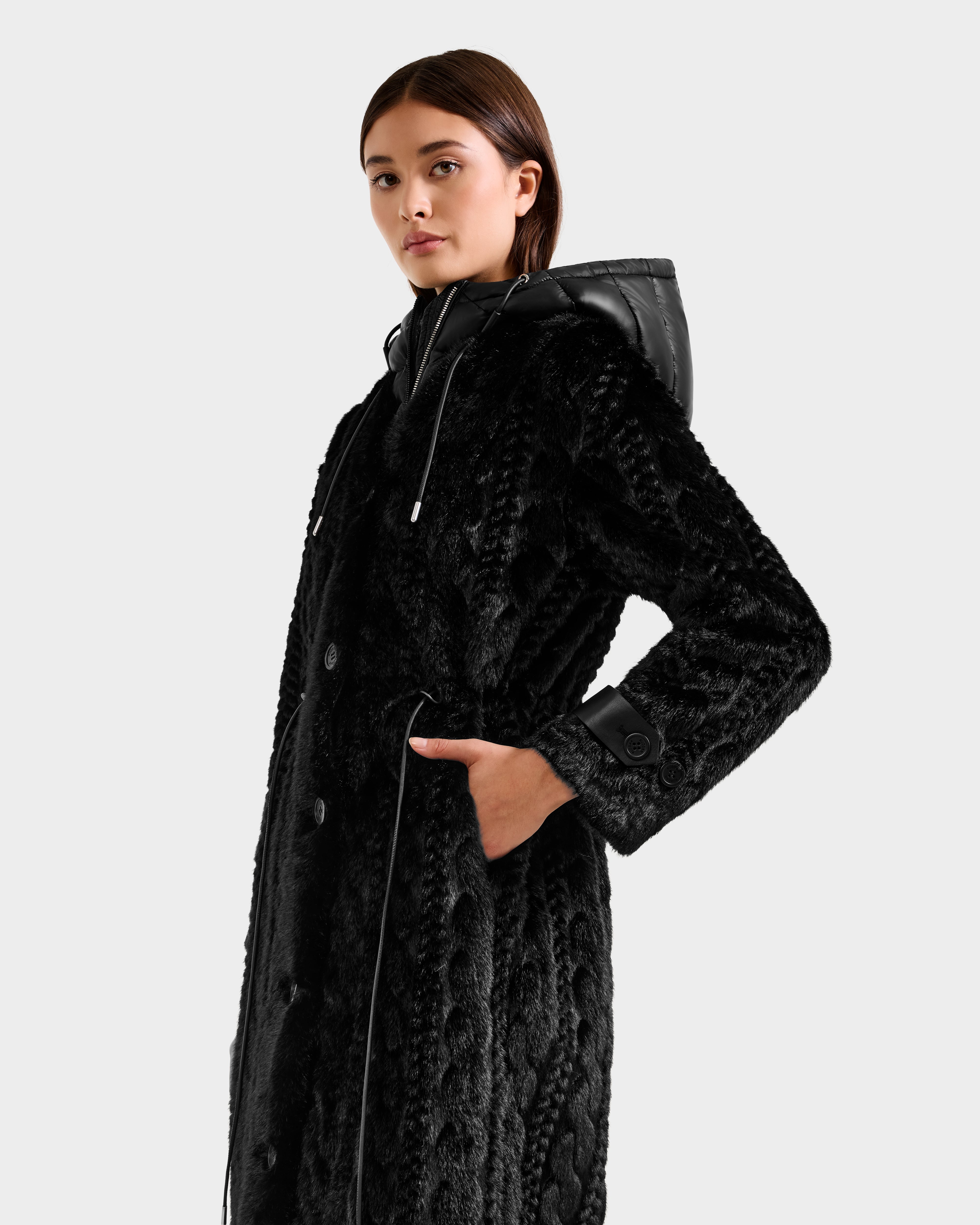 A woman stands confidently against a plain light background wearing the Rudsak ORIANA - 8125545 Black women's long textured faux fur coat with vegan leather trim and a quilted hood, one hand in her pocket.