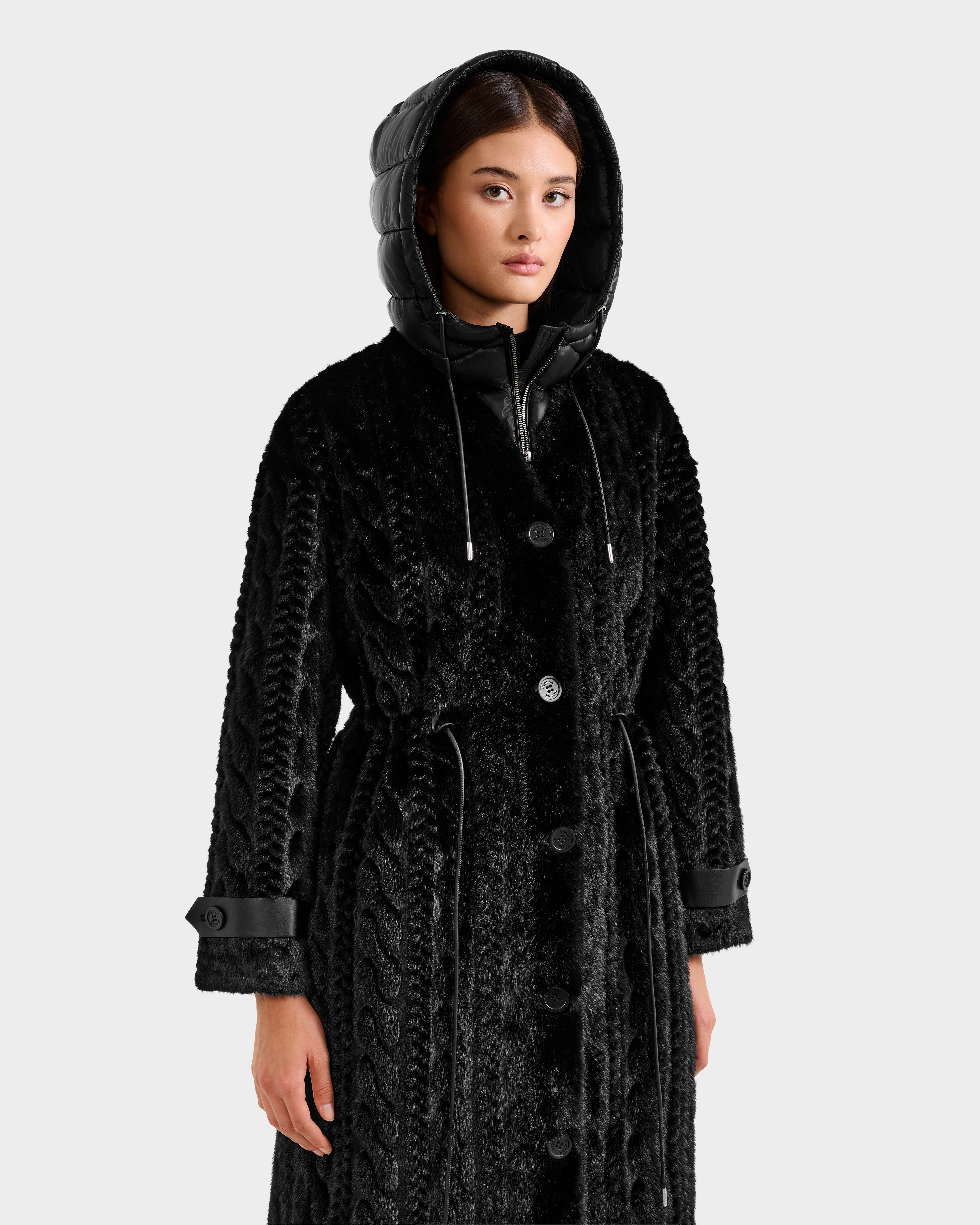 A woman models the Rudsak ORIANA - 8125545 Black coat, a textured black piece with a hood, buttoned front, drawstring waist, and vegan leather trims on the cuffs and hood, standing against a plain white background.
