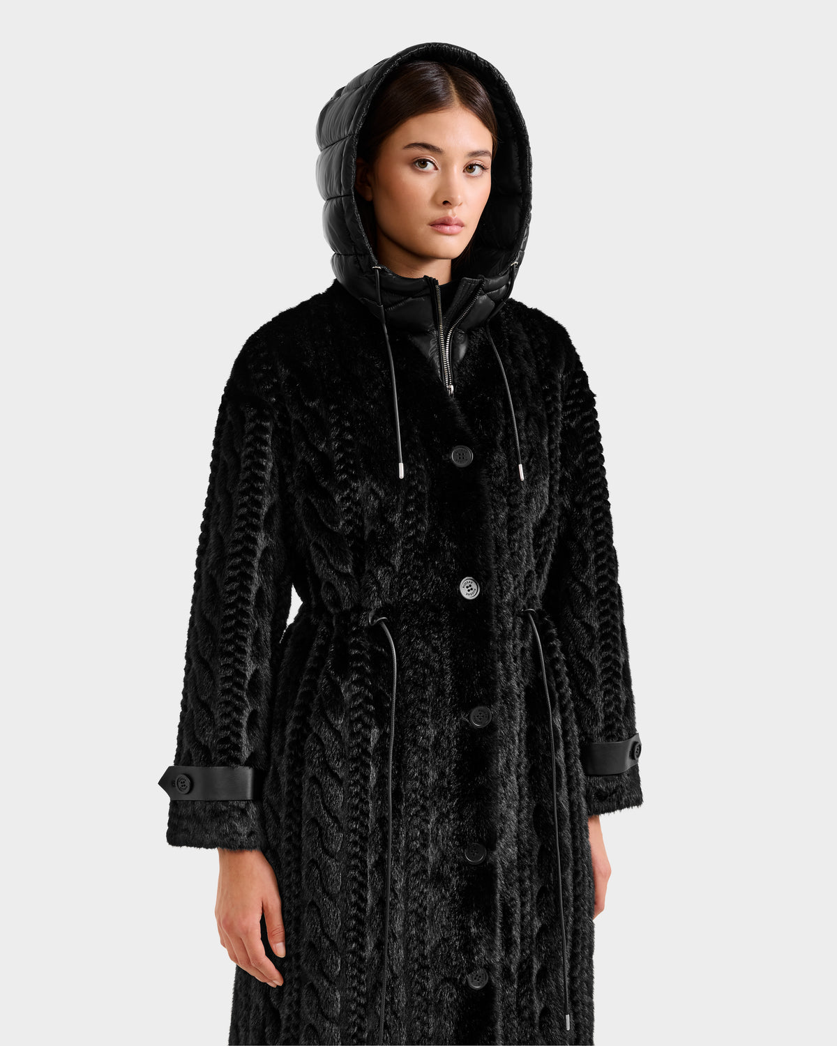 A woman models the Rudsak ORIANA - 8125545 Black coat, a textured black piece with a hood, buttoned front, drawstring waist, and vegan leather trims on the cuffs and hood, standing against a plain white background.