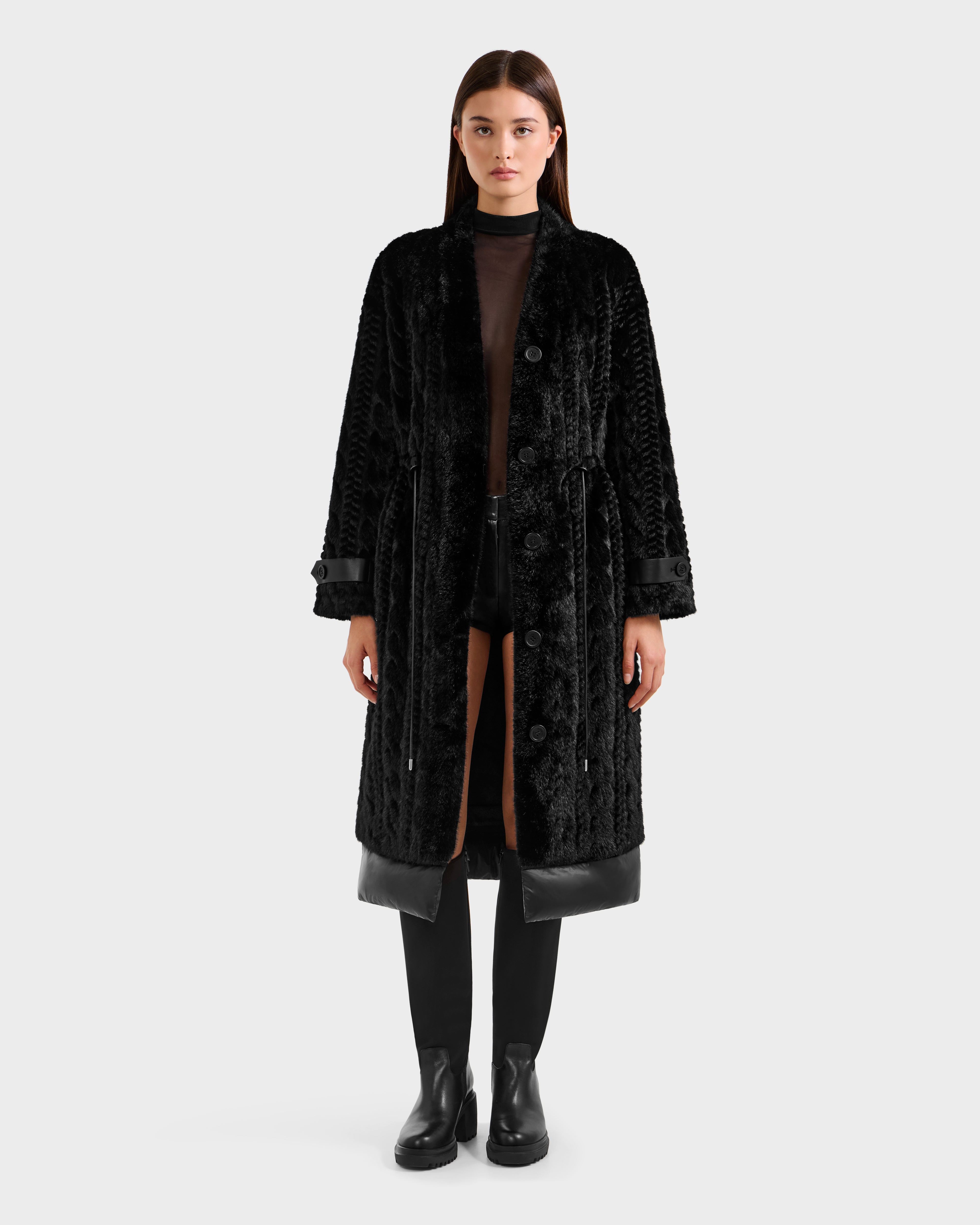 A woman faces forward wearing the Rudsak ORIANA - 8125545 Black long textured coat with vegan leather trims, styled with black boots and a sheer black top with shorts against a plain light gray background.